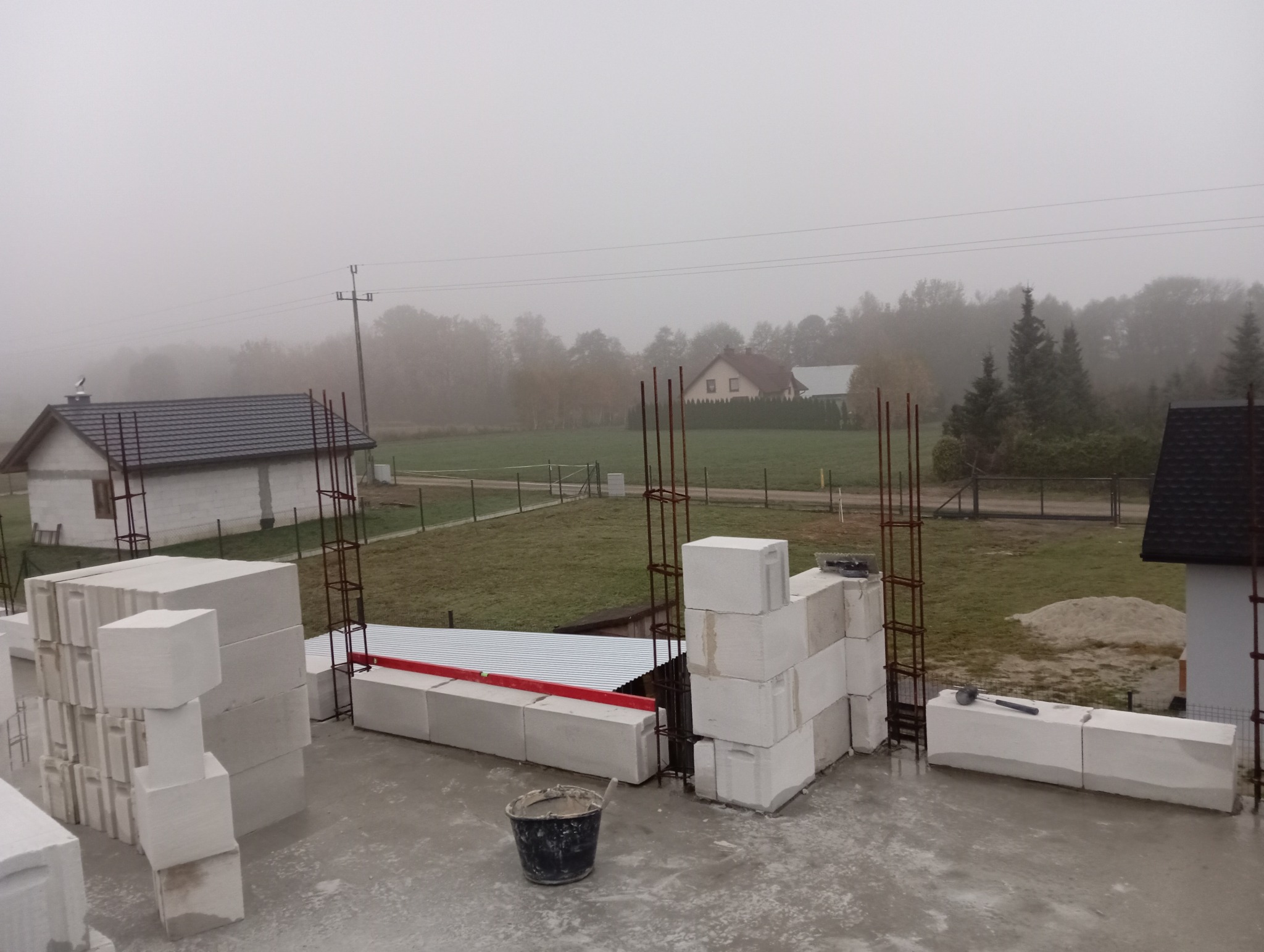 Budowa domu w trakcie: białe bloczki, zbrojenia, wiadro z zaprawą. W tle zabudowa jednorodzinna i mglista pogoda.