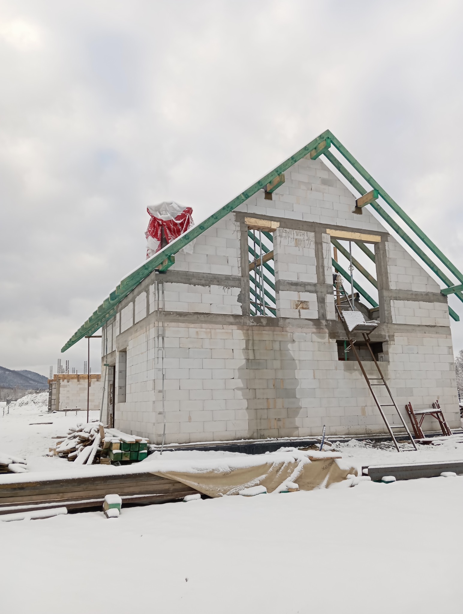 Surowy stan domu z białych bloczków w zimowej scenerii. Widoczne drewniane elementy konstrukcji dachu i rusztowanie. Częściowo ośnieżone materiały budowlane na pierwszym planie.