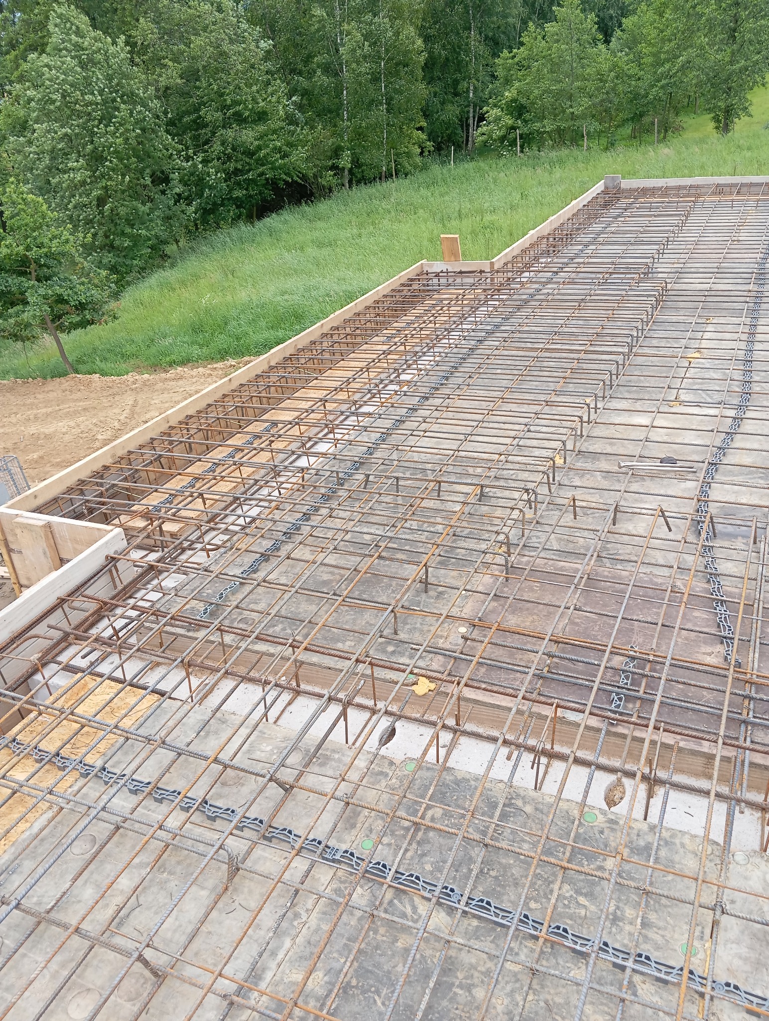 Zbrojenie płyty fundamentowej z użyciem dystansów, widok z góry na tle zieleni. Przygotowanie do wylewania betonu, etap budowy domu.
