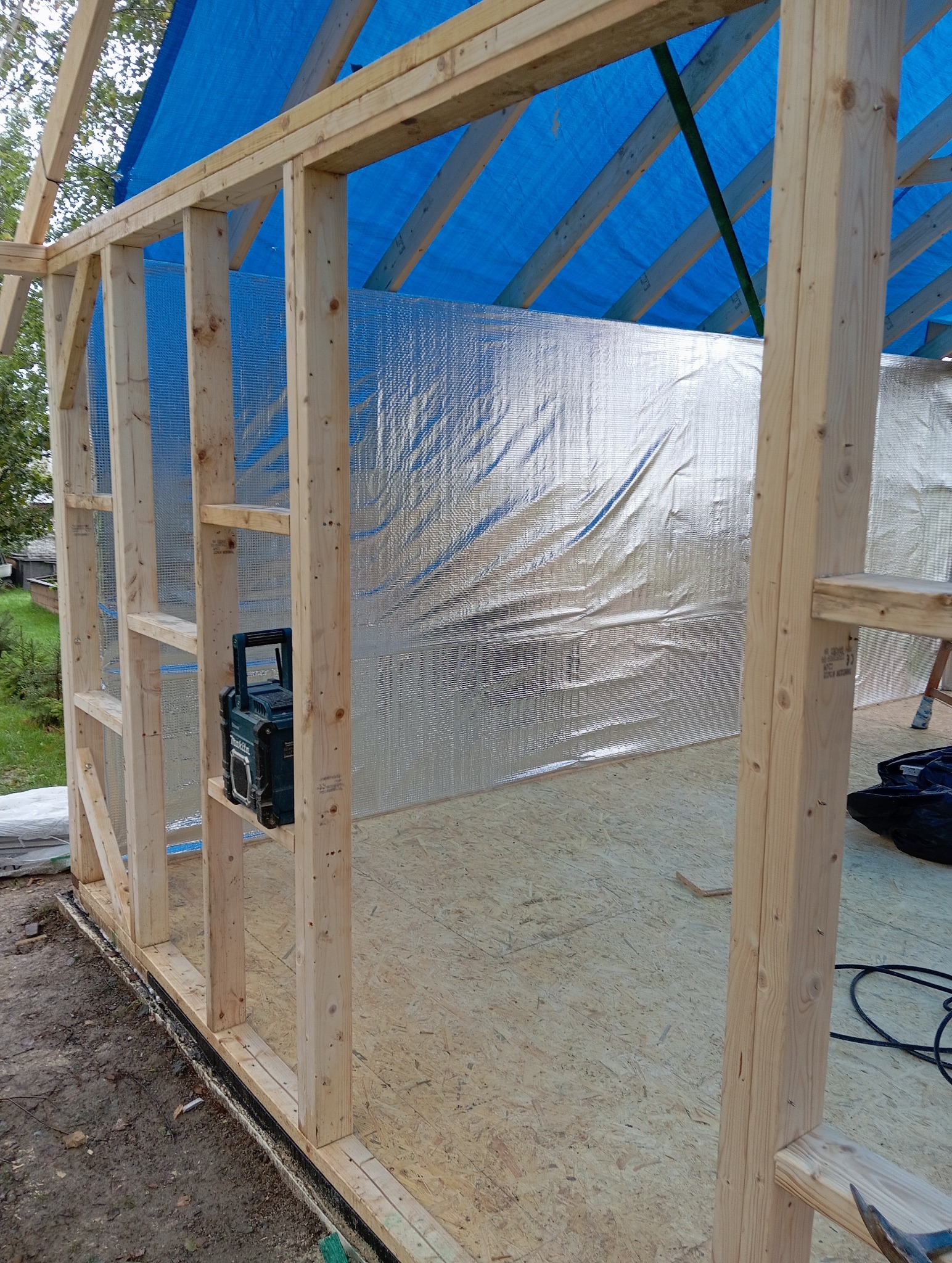 Wnętrze szkieletu domu w budowie z izolacją termiczną i niebieską plandeką na dachu. Widoczny radiobudzik na drewnianej konstrukcji.