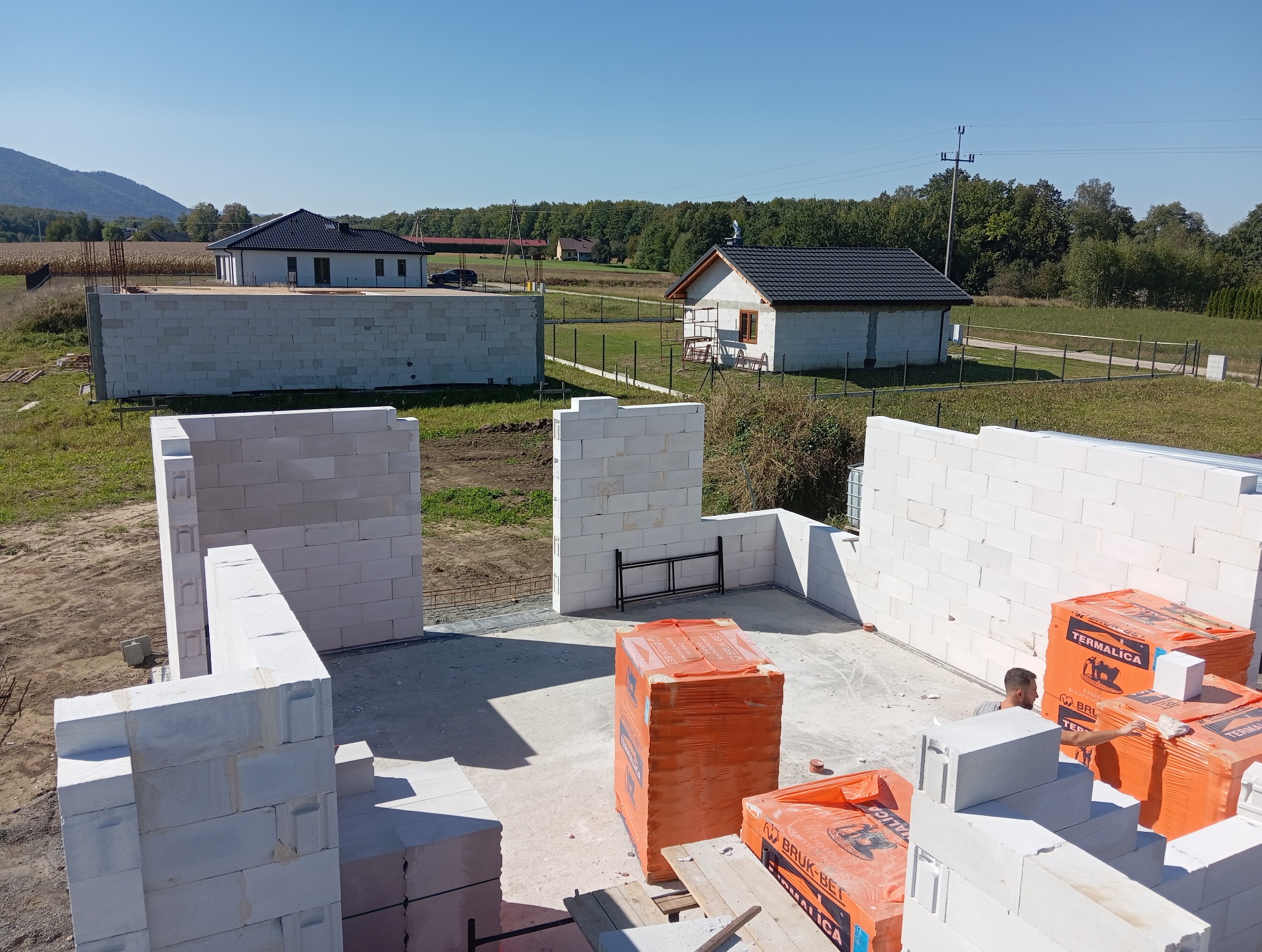 Budowa domu w toku: wznoszone ściany z białych bloczków, palety z materiałami budowlanymi. W tle dwa ukończone domy i zielone wzgórza.