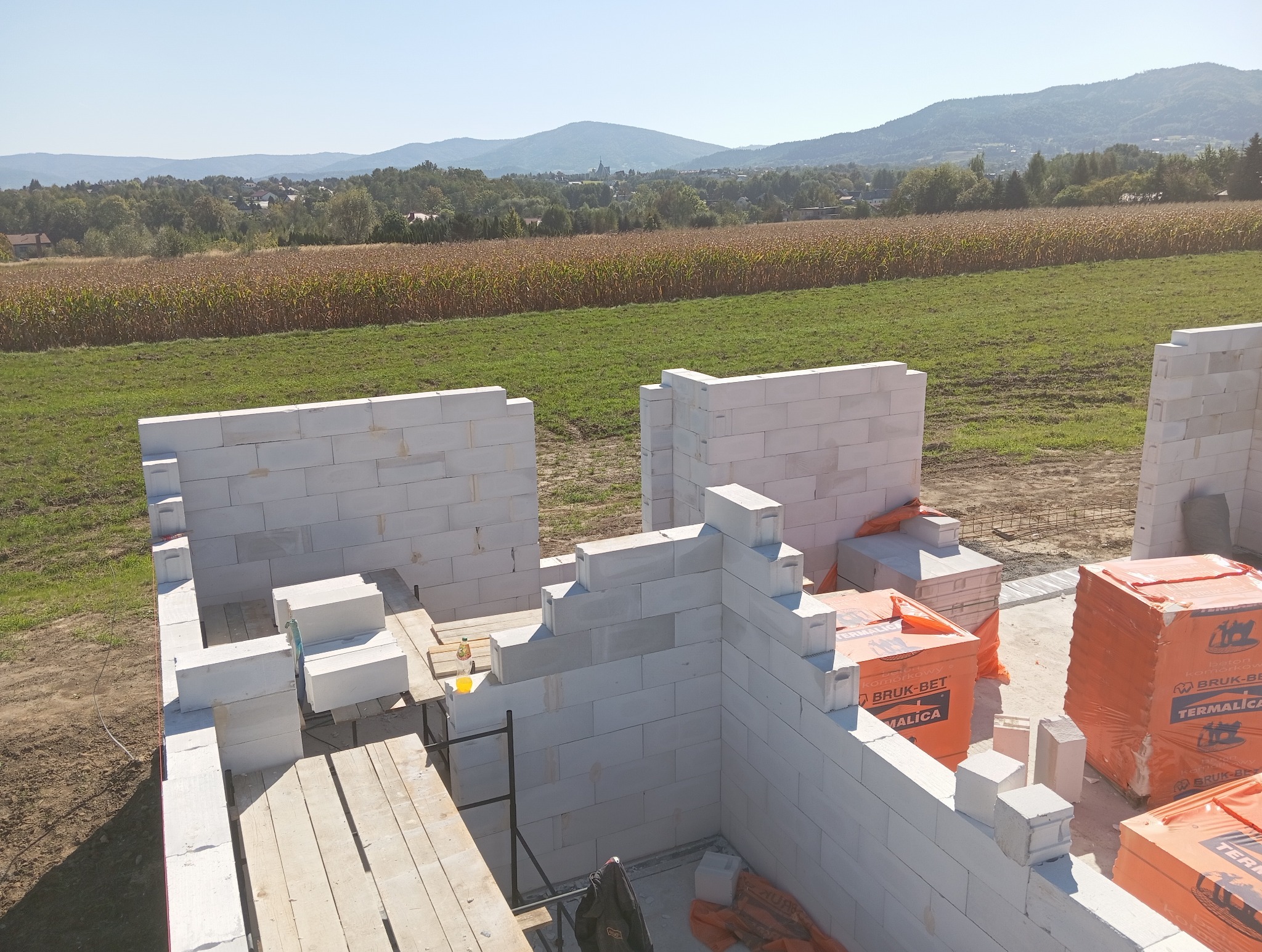 Wznoszenie ścian domu z bloczków betonu komórkowego na tle krajobrazu z polami i wzgórzami. Częściowo wymurowane ściany, palety z bloczkami.