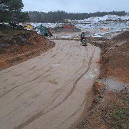 Seweryn Drewa - Prace ziemne: zagęszczanie podłoża pod drogę z użyciem zagęszczarki w kopalni piasku. W tle maszyny górnicze i hałdy piasku. Pochmurny dzień.