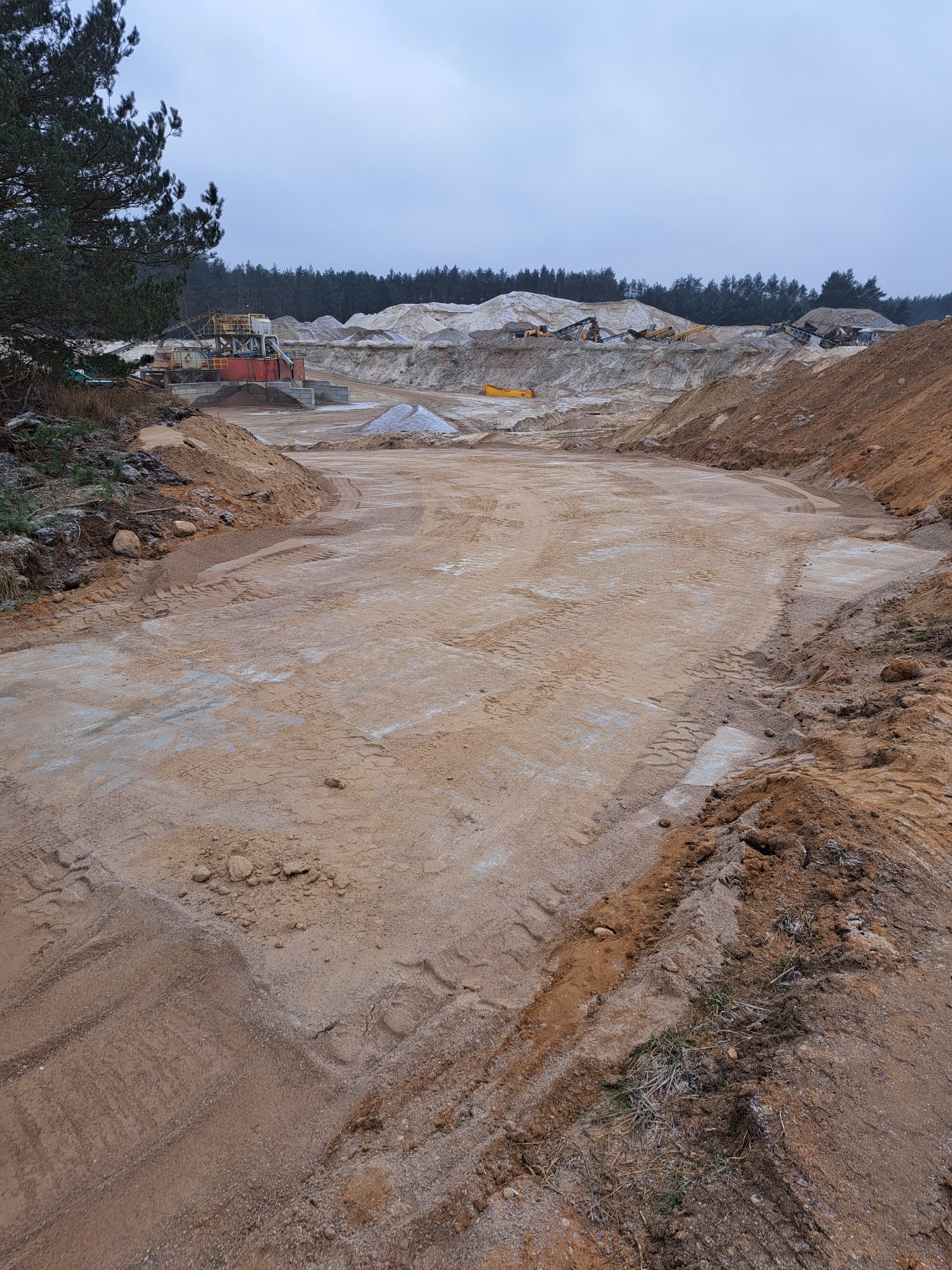 Wyrównany teren piaskowy z widocznymi śladami opon, w tle maszyny budowlane i hałdy piasku. Pochmurne niebo. Prace ziemne w toku.