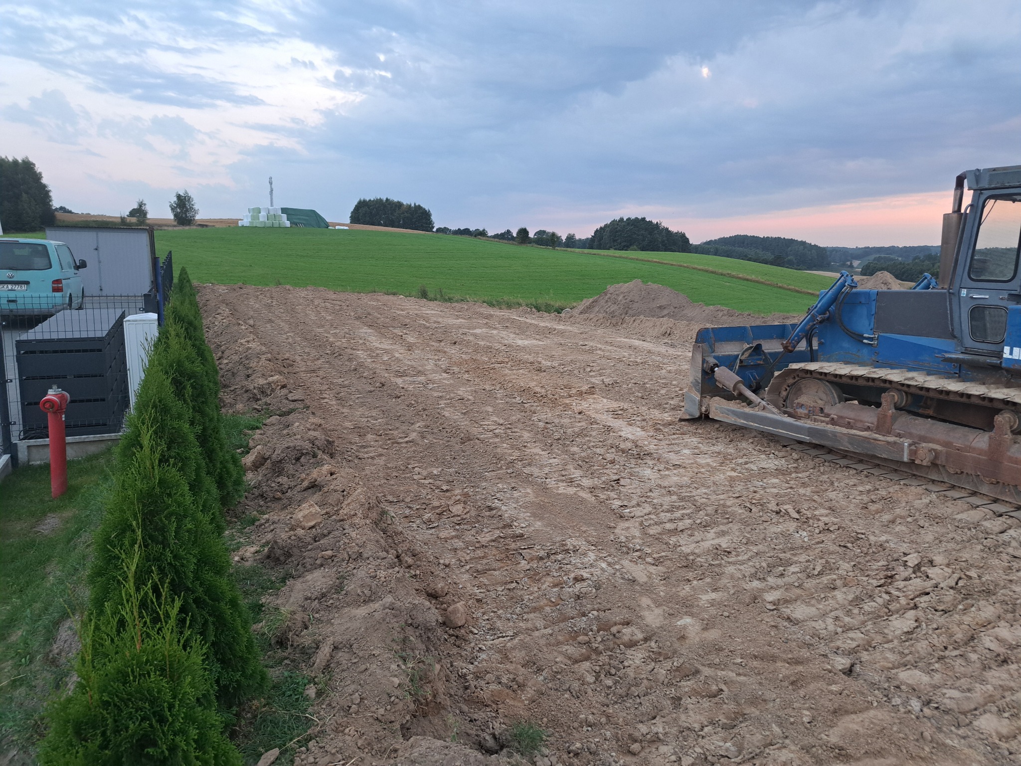 Niebieski spychacz wyrównuje teren pod budowę, widok na zielone pola i zachmurzone niebo. Ślady gąsienic na ziemi, rząd tuj wzdłuż ogrodzenia.