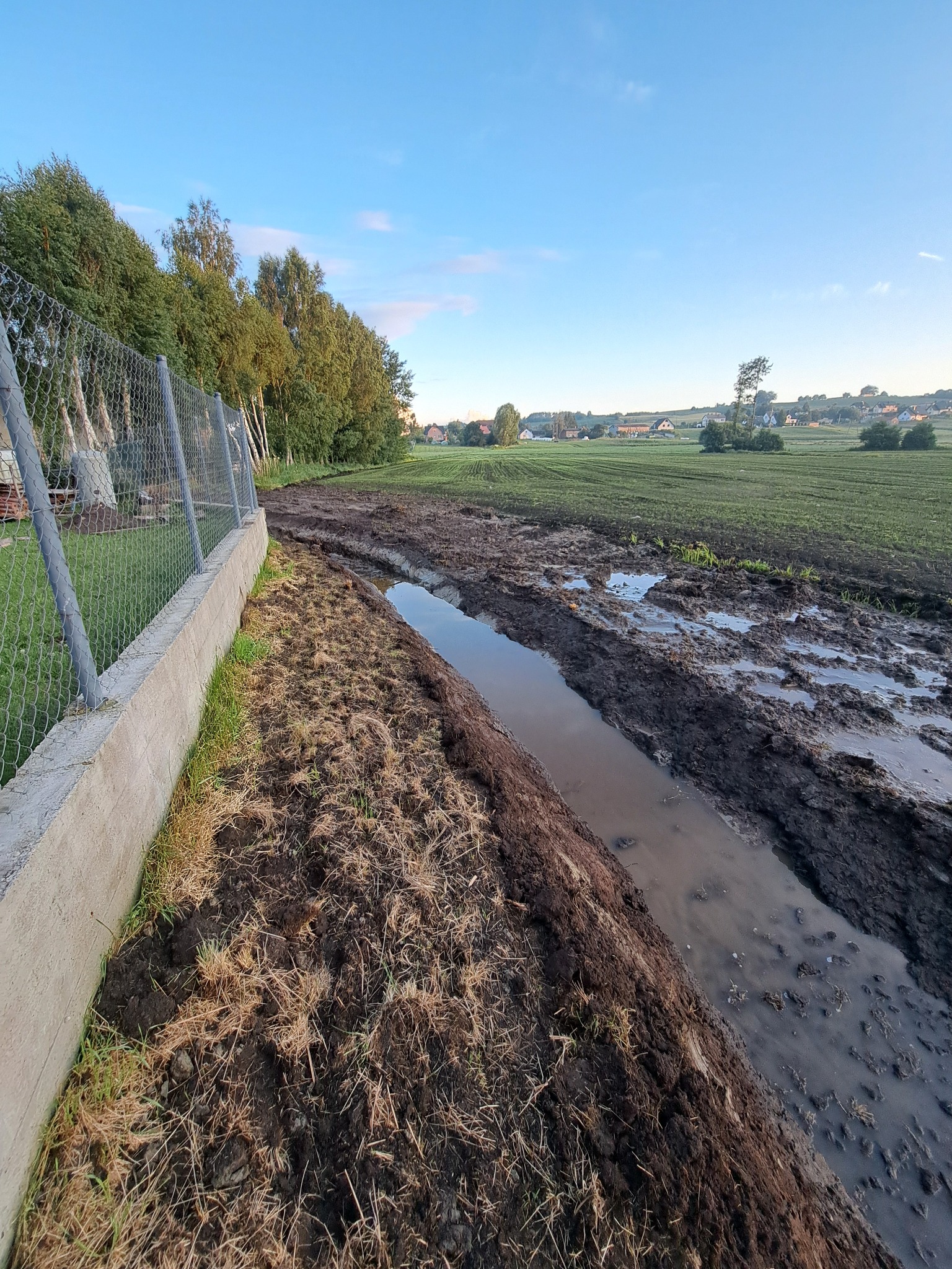 Świeżo wykopany rów melioracyjny wzdłuż ogrodzenia z siatki, z widocznym błotnistym terenem i polami w tle, w słoneczny dzień. Prace ziemne.