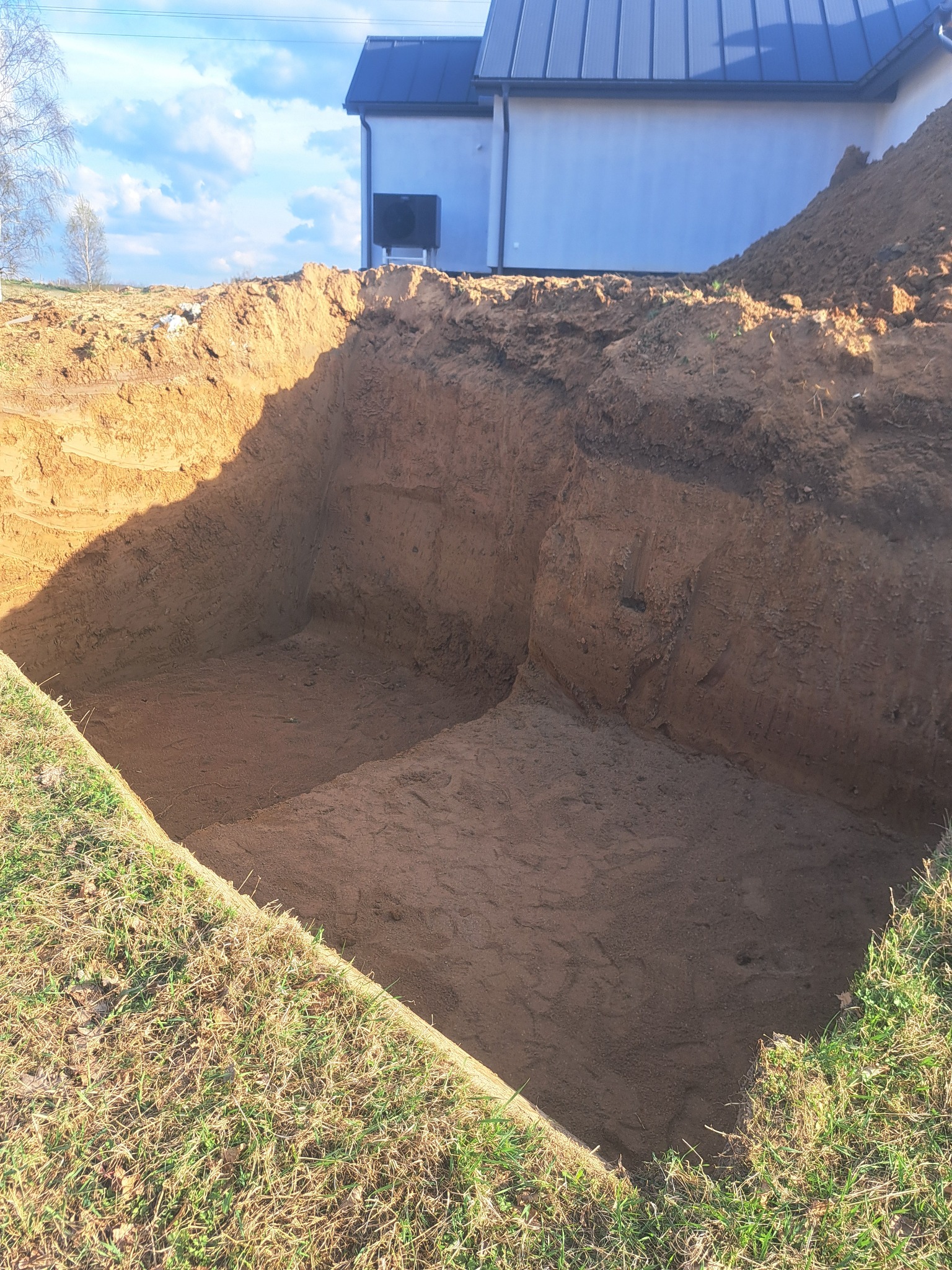 Wykopany, kwadratowy dół w ziemi, prawdopodobnie pod fundamenty, z widocznym budynkiem w tle i stertą ziemi obok. Świeżo wykopana ziemia.