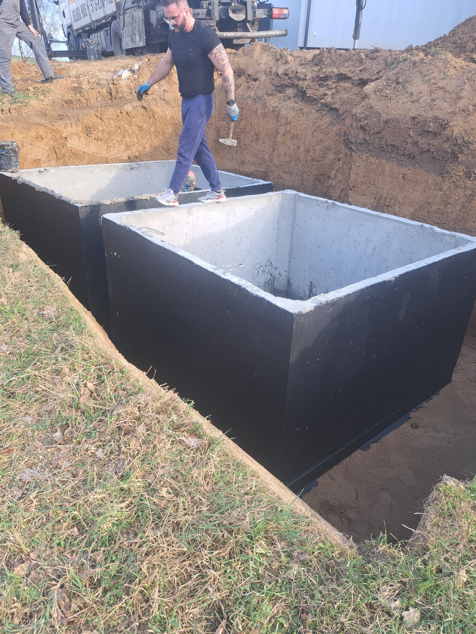 Dwa betonowe zbiorniki w wykopie, zabezpieczone czarną powłoką. Mężczyzna w okularach przechodzi po krawędzi jednego z nich. Widoczny fragment ciężarówki w tle.