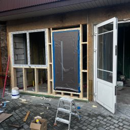 Yszbud - Montaż stolarki okiennej i drzwiowej w surowym stanie. Widoczne drewniane ramy, okno z szybami i drzwi zabezpieczone folią. Na podłożu narzędzia i resztki materiałów.