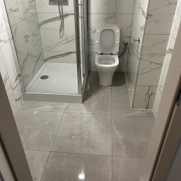 Yszbud - Wykończona łazienka z kabiną prysznicową i toaletą, płytki imitujące marmur na ścianach i podłodze, nowoczesny design, widok zza framugi drzwi.