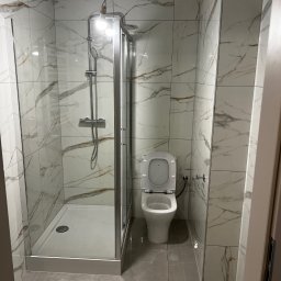 Yszbud - Wykończona łazienka z kabiną prysznicową i toaletą. Białe płytki imitujące marmur z brązowymi żyłami. Szklana kabina prysznicowa z chromowanym wykończeniem.