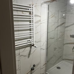 Yszbud - Biała łazienka z marmurowymi płytkami, grzejnikiem drabinkowym i kabiną prysznicową z przezroczystego szkła. Minimalistyczny design, nowoczesne wykończenie.