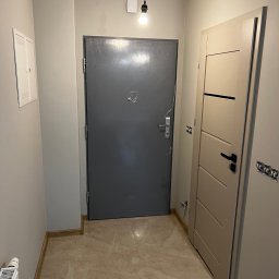Yszbud - Korytarz po remoncie: szare drzwi wejściowe, beżowe drzwi wewnętrzne z czarną klamką, marmurowe płytki podłogowe i świeżo pomalowane ściany w odcieniach szarości.