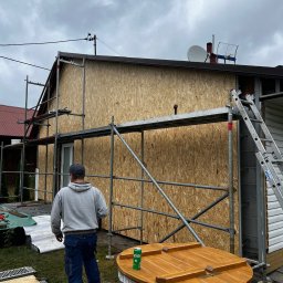 Yszbud - Dom w trakcie ocieplania: ściany obłożone płytami OSB, rusztowania, drabina. Widoczny fragment ogrodu z drewnianą studnią. Pochmurne niebo w tle.