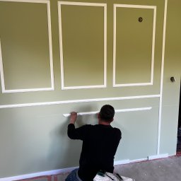 Yszbud - Pracownik montuje białe listwy dekoracyjne na zielonej ścianie z panelami. Widoczny drabinka i narzędzia. Detal wykończenia wnętrza w stylu klasycznym.