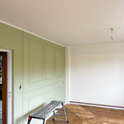 Yszbud - Pokój po malowaniu: ściana w odcieniach zieleni z dekoracyjnymi panelami, biała ściana, drabina malarska, parkiet. Widoczna osoba w tle.