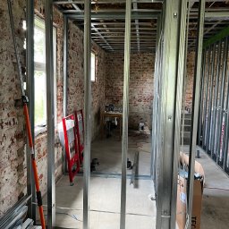 Yszbud - Montaż metalowej konstrukcji ścian działowych w surowym wnętrzu z ceglanymi ścianami. Widoczne profile, narzędzia i elementy instalacji. Prace wykończeniowe w trakcie.