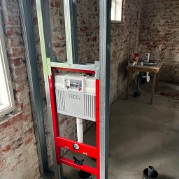 Yszbud - Stelaż podtynkowy TECE zamontowany na metalowej konstrukcji w surowym wnętrzu z ceglaną ścianą. Widoczna instalacja wodno-kanalizacyjna i okno w tle.