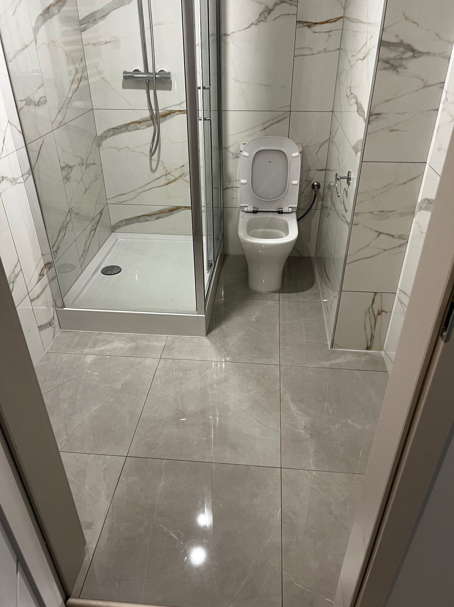 Wykończona łazienka z kabiną prysznicową i toaletą, płytki imitujące marmur na ścianach i podłodze, nowoczesny design, widok zza framugi drzwi.