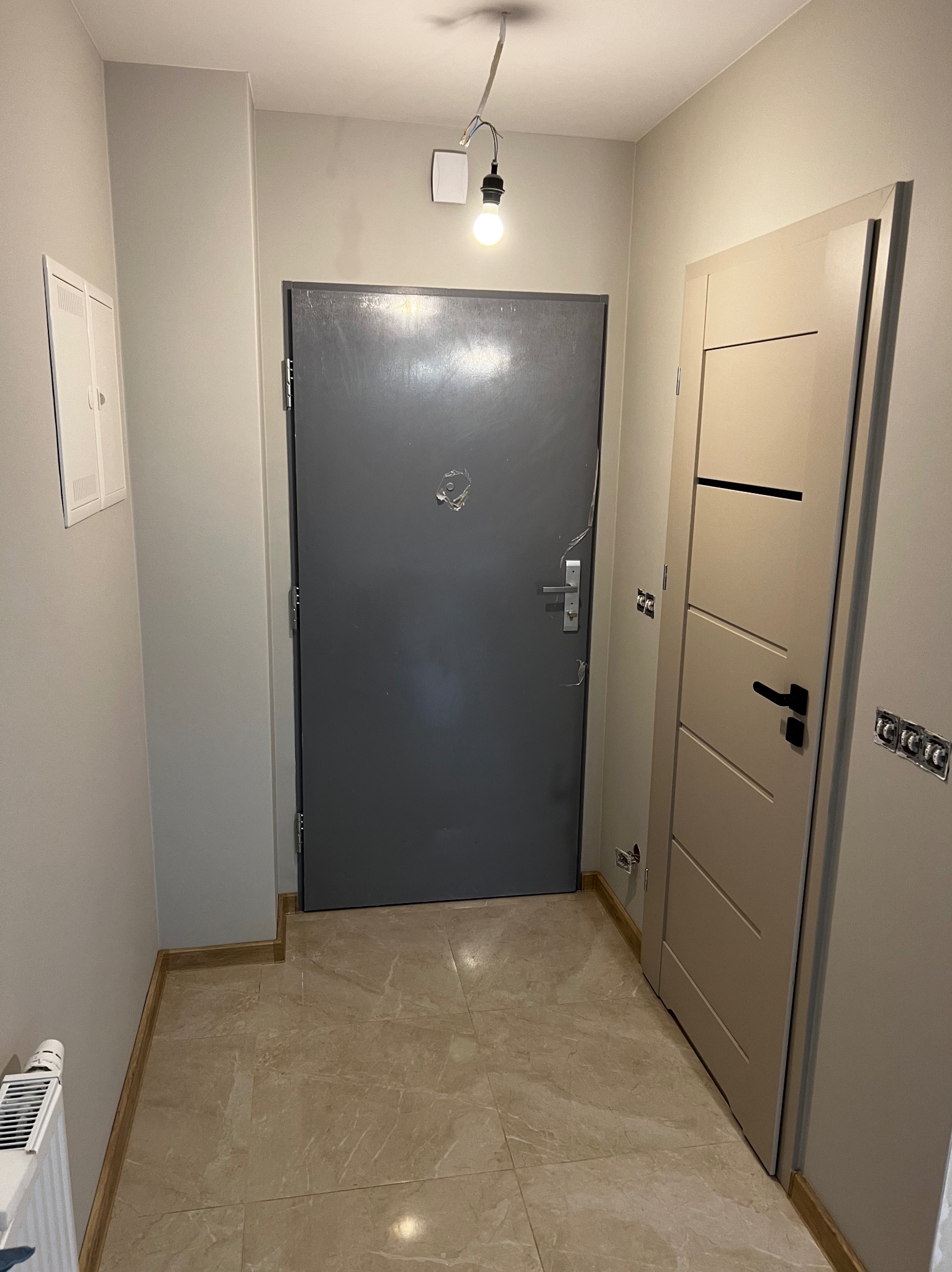 Korytarz po remoncie: szare drzwi wejściowe, beżowe drzwi wewnętrzne z czarną klamką, marmurowe płytki podłogowe i świeżo pomalowane ściany w odcieniach szarości.