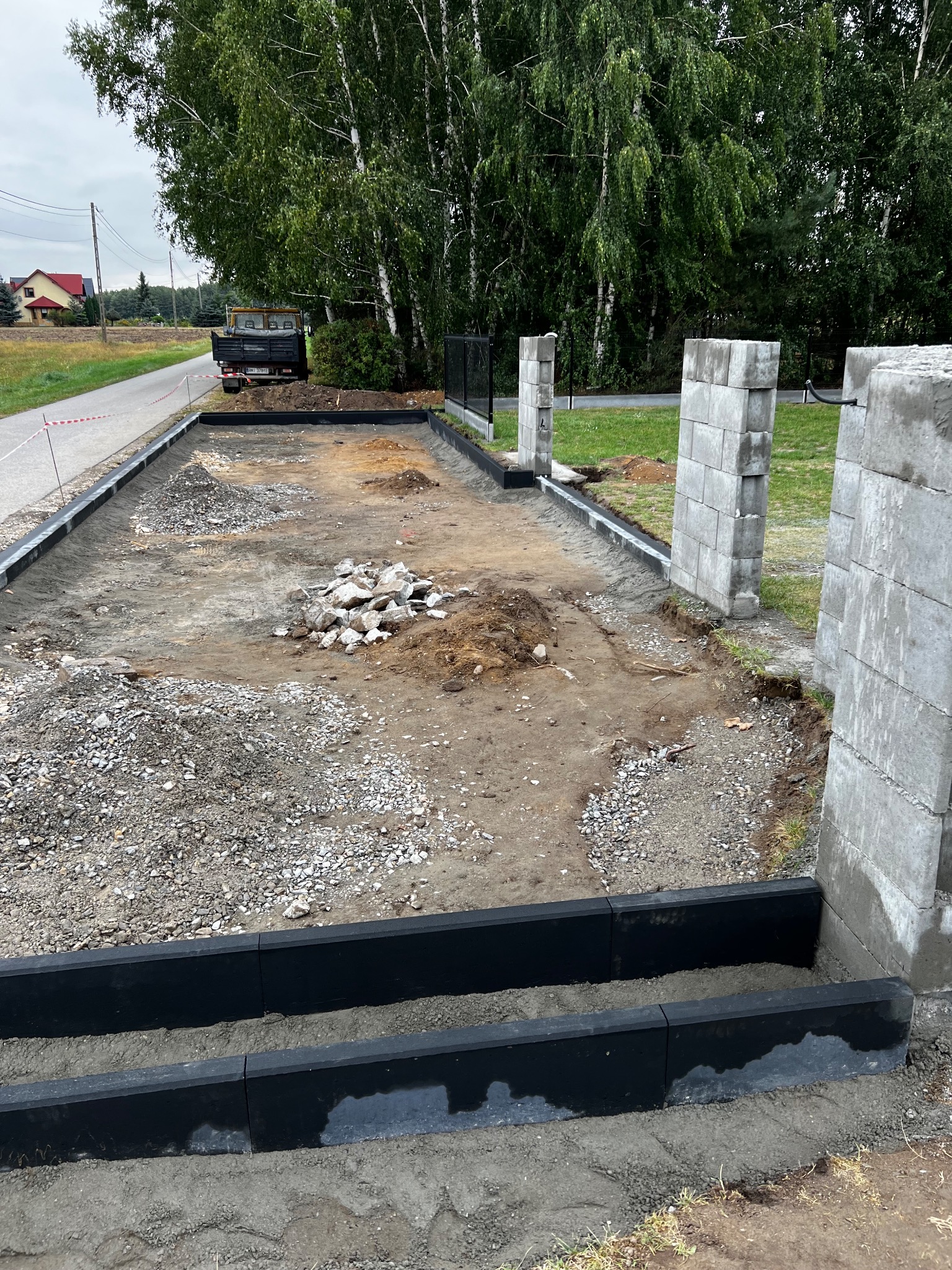 Przygotowanie terenu pod budowę ogrodzenia: ułożone obrzeża, fundamenty słupków z bloczków betonowych, gruz i żwir na ziemi. W tle drzewa i budynek.