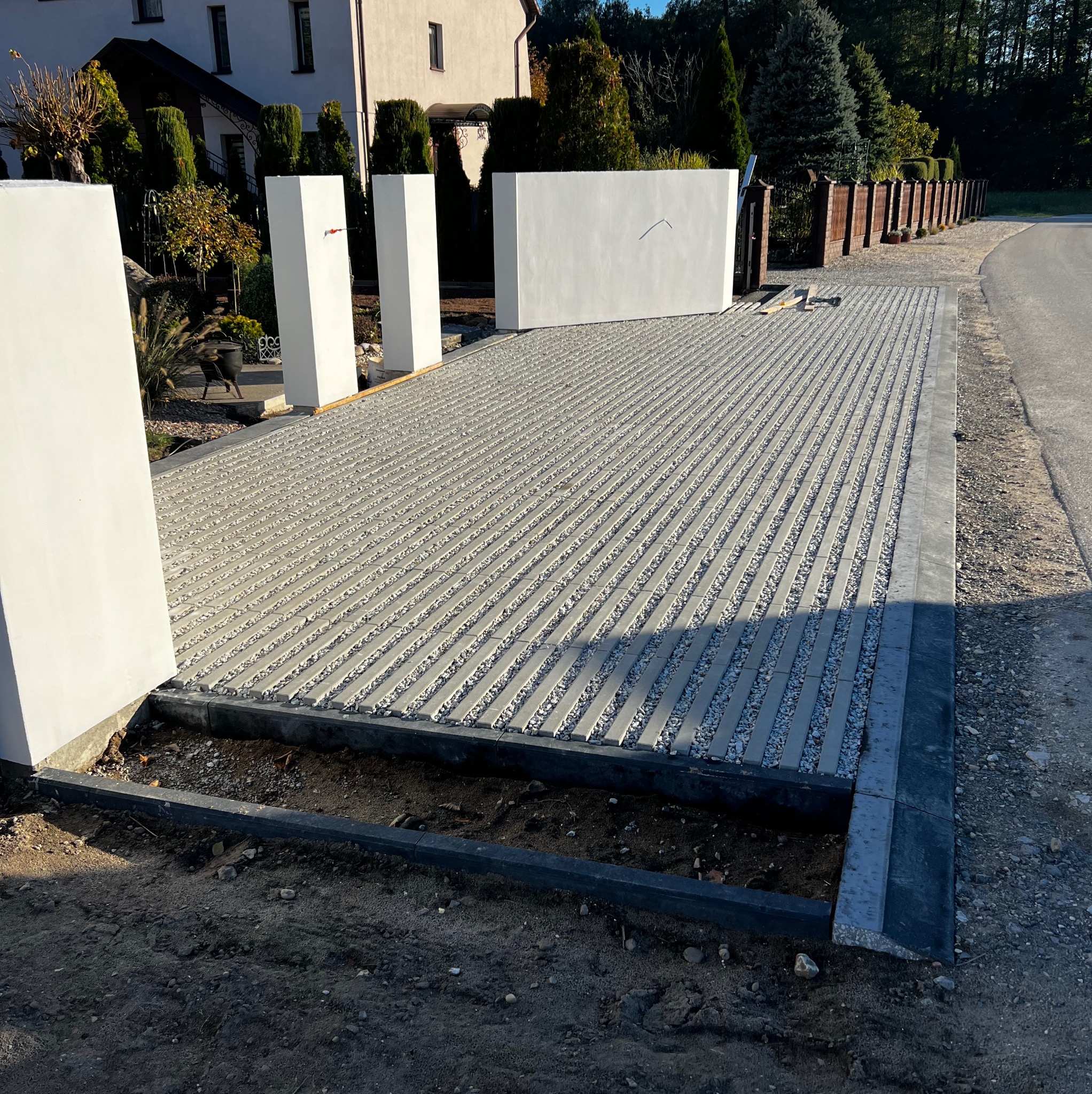 Układanie nawierzchni z ażurowych płyt betonowych wypełnionych grysem na podjeździe. Widoczne białe słupki ogrodzeniowe i fragment domu w tle.