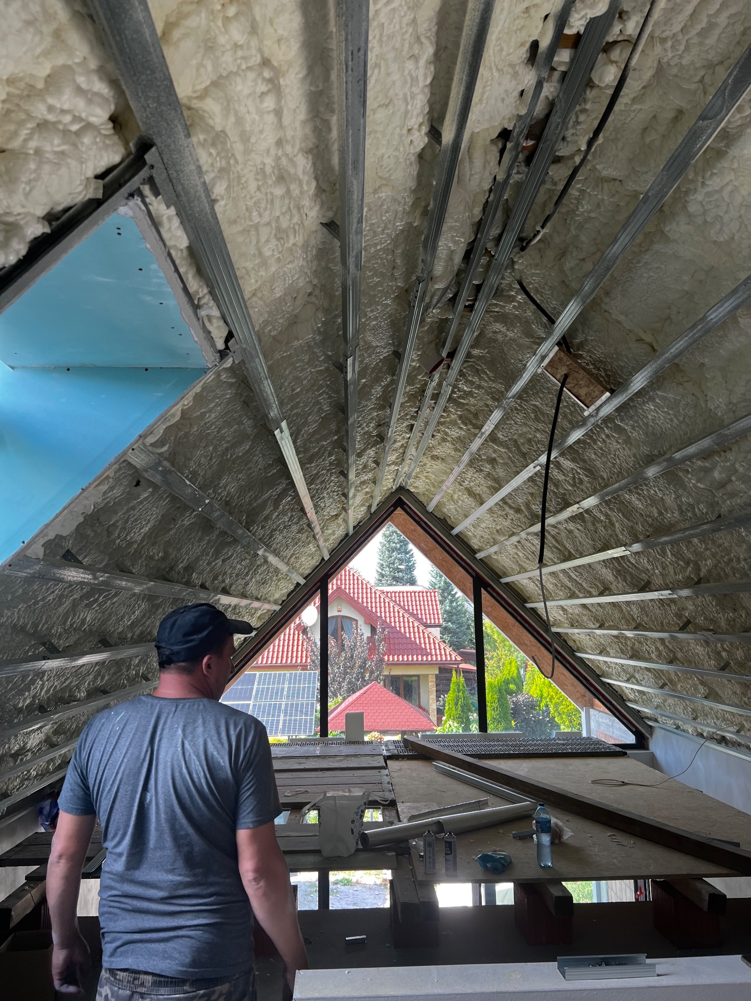 Ocieplenie poddasza pianą PUR z widoczną konstrukcją metalową i fragmentem płyty gipsowej. W tle widok na dom przez okno w szczycie dachu.