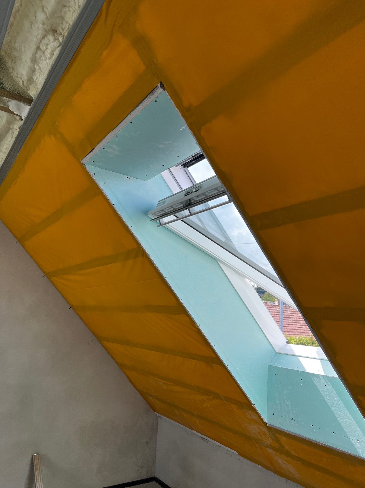 Montaż okna dachowego w skośnej ścianie poddasza, widoczne płyty karton-gips i folia paroizolacyjna. Ujęcie z dołu, okno otwarte, widok na niebo i dach budynku.