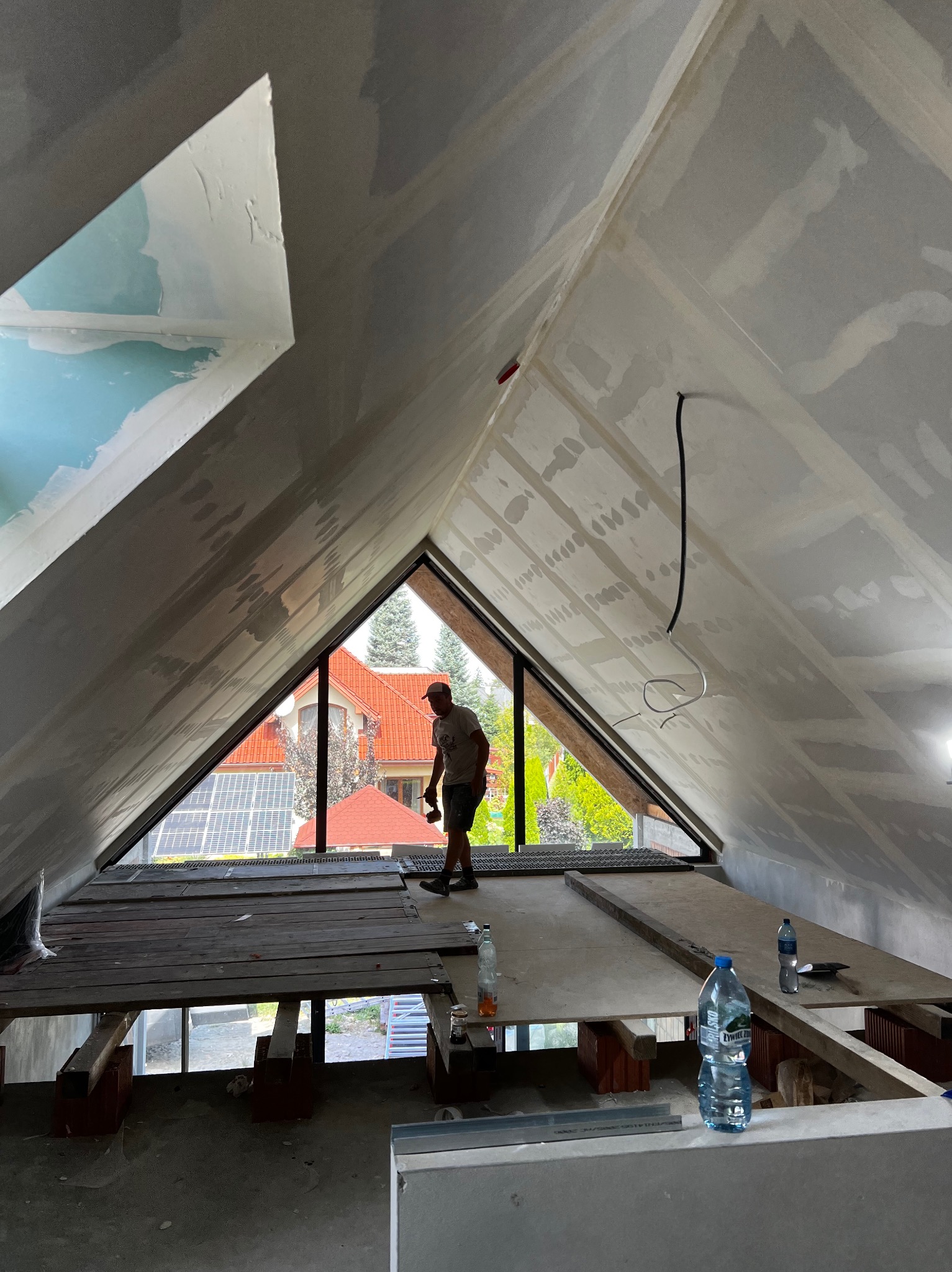 Adaptacja poddasza: mężczyzna montuje płyty karton-gips na skośnym suficie, widoczne okno z widokiem na dom z czerwoną dachówką i panele słoneczne. Prace wykończeniowe.