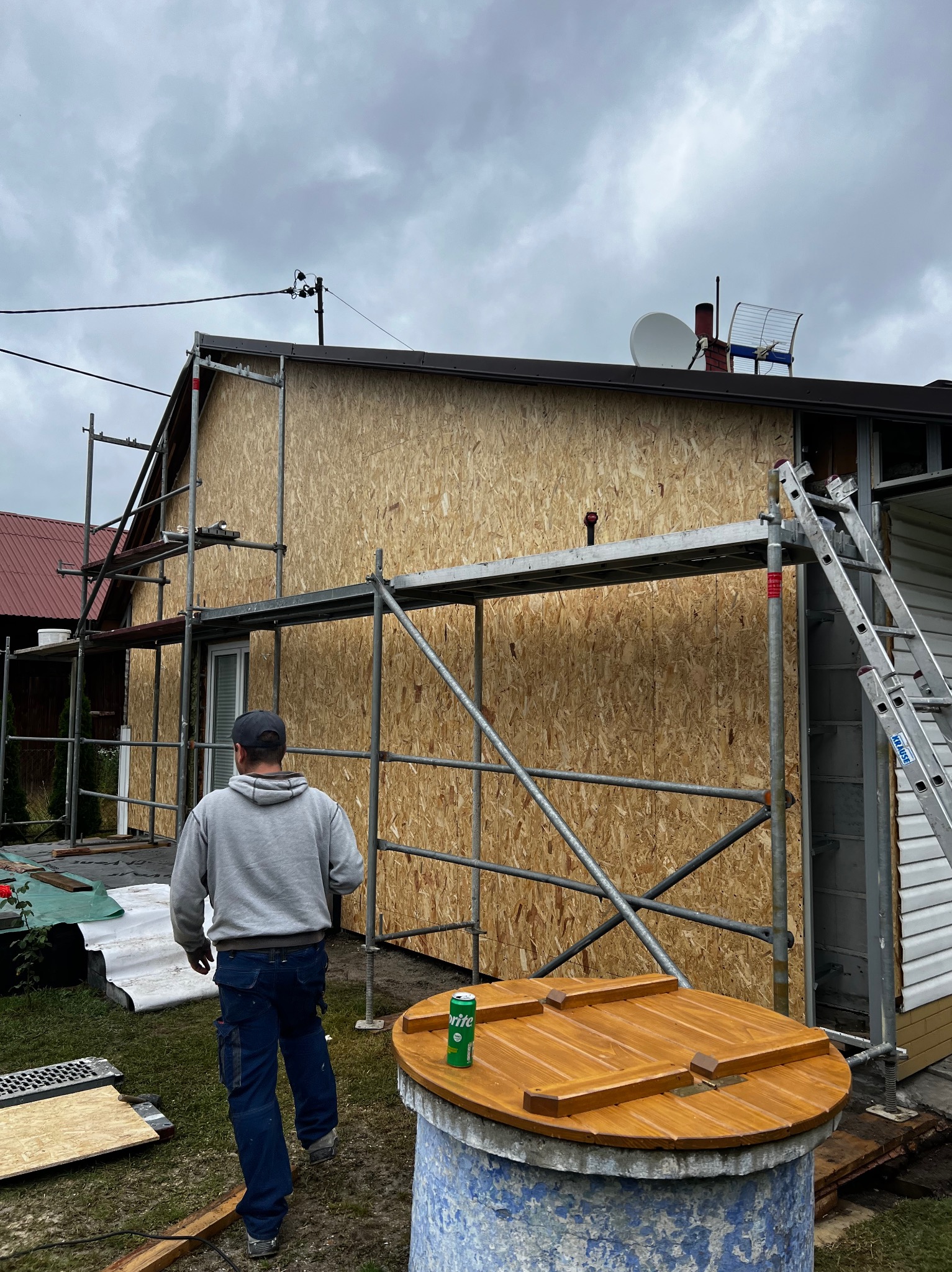 Dom w trakcie ocieplania: ściany obłożone płytami OSB, rusztowania, drabina. Widoczny fragment ogrodu z drewnianą studnią. Pochmurne niebo w tle.
