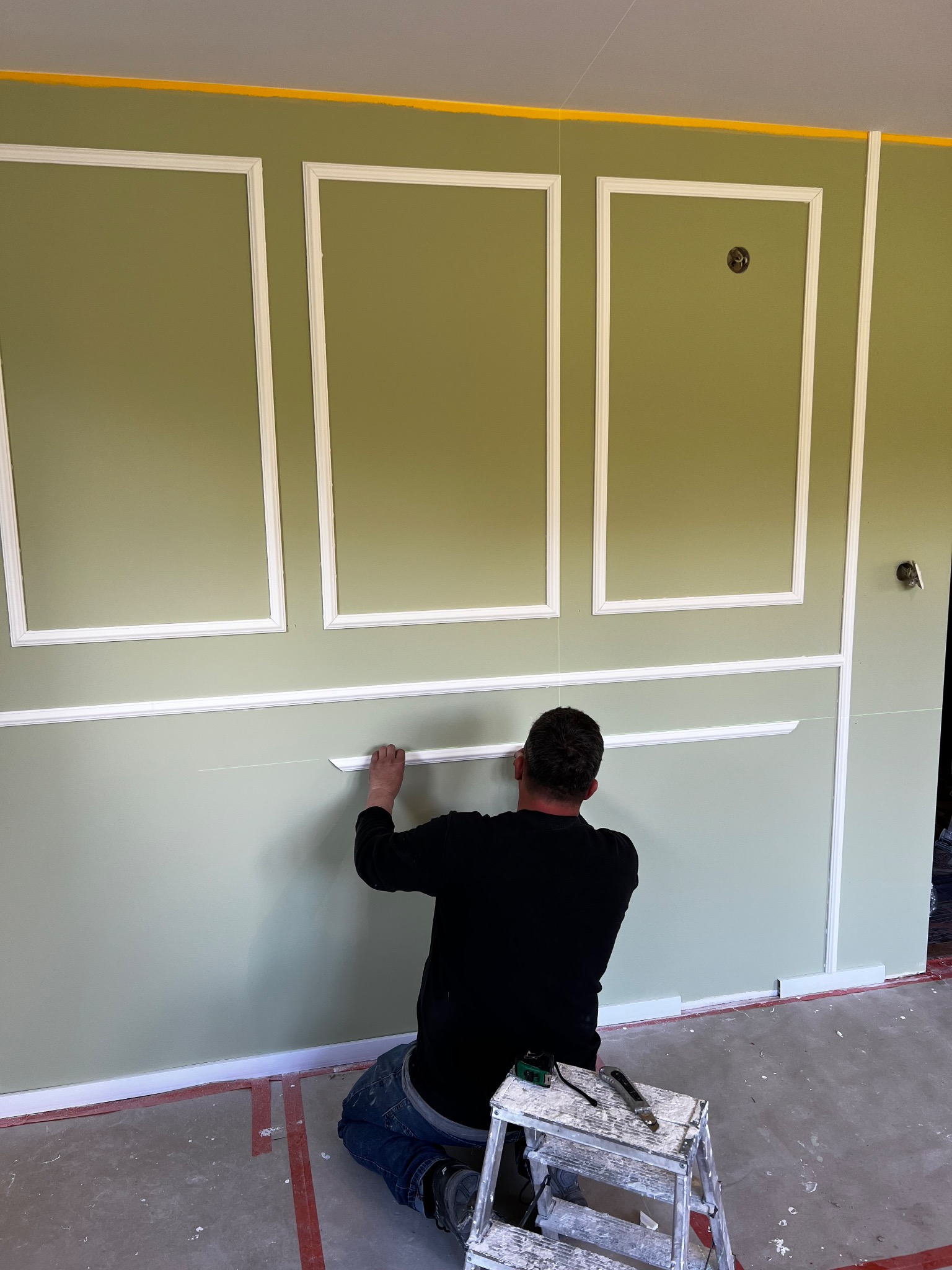 Pracownik montuje białe listwy dekoracyjne na zielonej ścianie z panelami. Widoczny drabinka i narzędzia. Detal wykończenia wnętrza w stylu klasycznym.