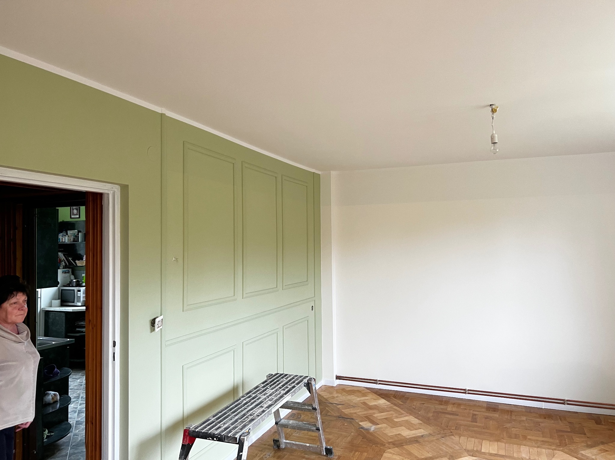 Pokój po malowaniu: ściana w odcieniach zieleni z dekoracyjnymi panelami, biała ściana, drabina malarska, parkiet. Widoczna osoba w tle.