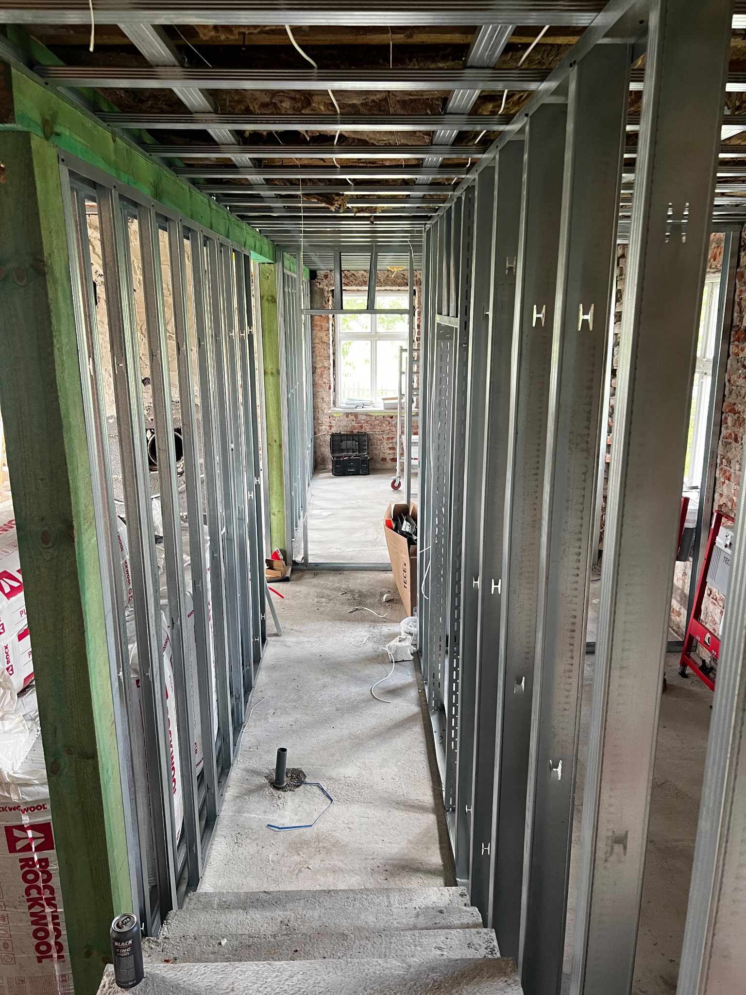 Metalowa konstrukcja ścian działowych w surowym wnętrzu, widok wzdłuż korytarza. Widoczne profile, izolacja Rockwool i fragmenty instalacji. Etap prac wykończeniowych.