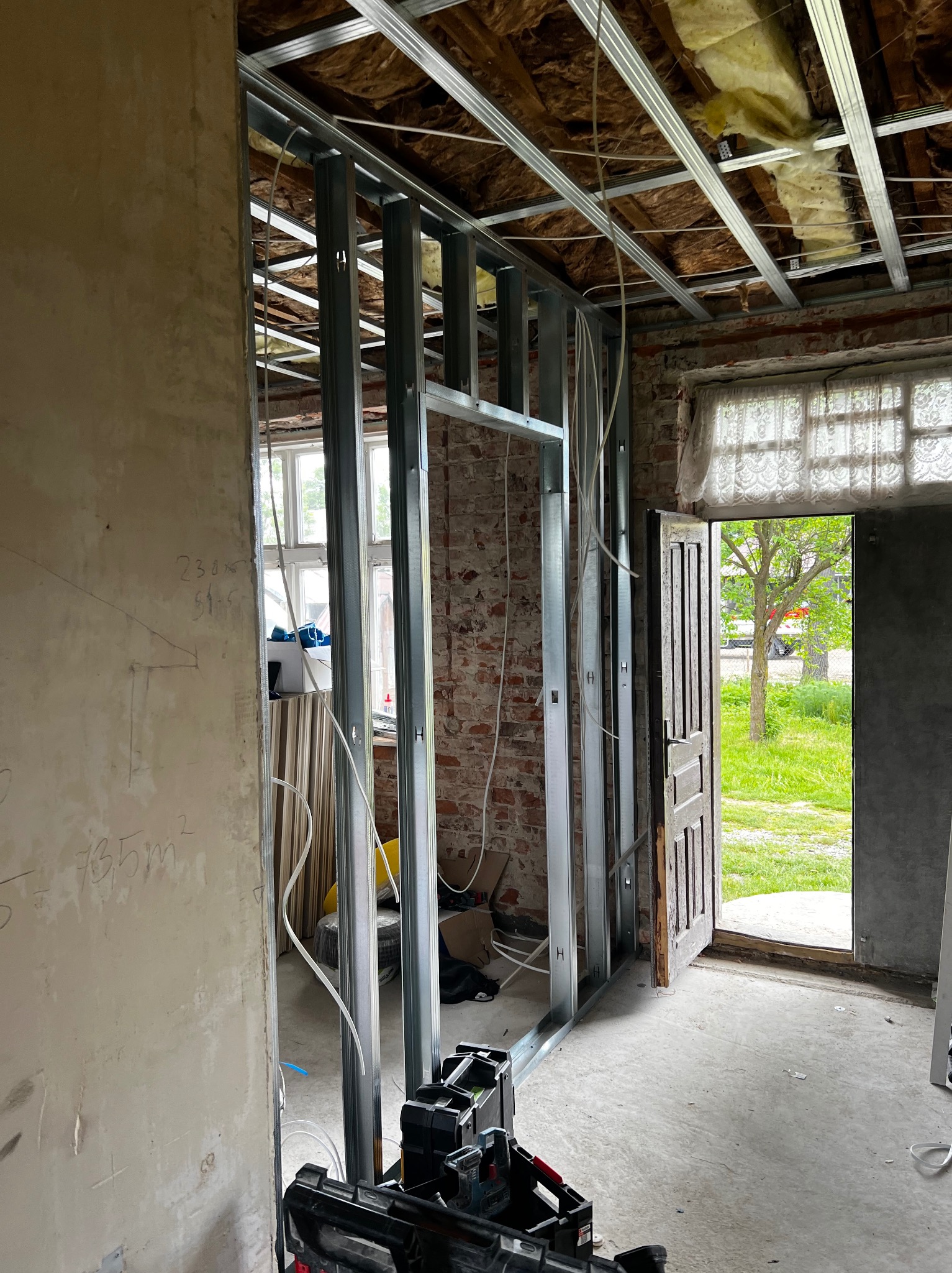 Metalowa konstrukcja ścianki działowej z płyt karton-gips, widoczne przewody elektryczne, surowa cegła i narzędzia. W tle otwarte drzwi z widokiem na zieleń.