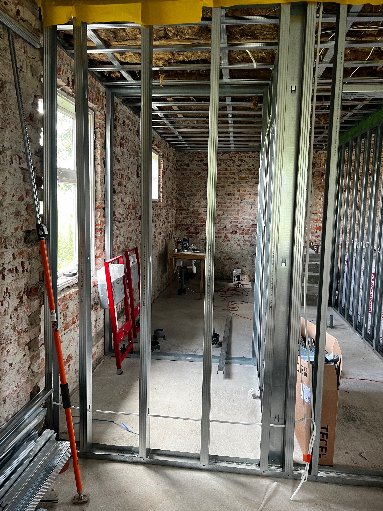 Montaż metalowej konstrukcji ścian działowych w surowym wnętrzu z ceglanymi ścianami. Widoczne profile, narzędzia i elementy instalacji. Prace wykończeniowe w trakcie.