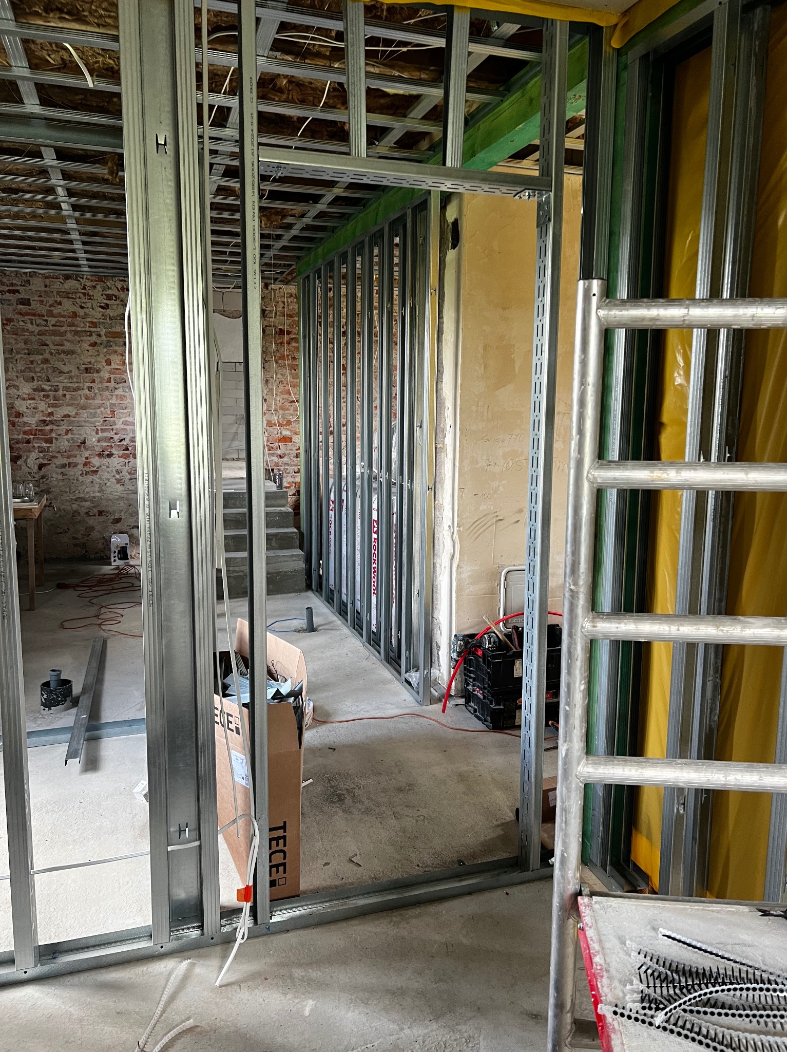 Metalowa konstrukcja ścian działowych z płyt karton-gips, widok na surową cegłę i schody w tle. Etap budowy z widocznymi narzędziami i materiałami.