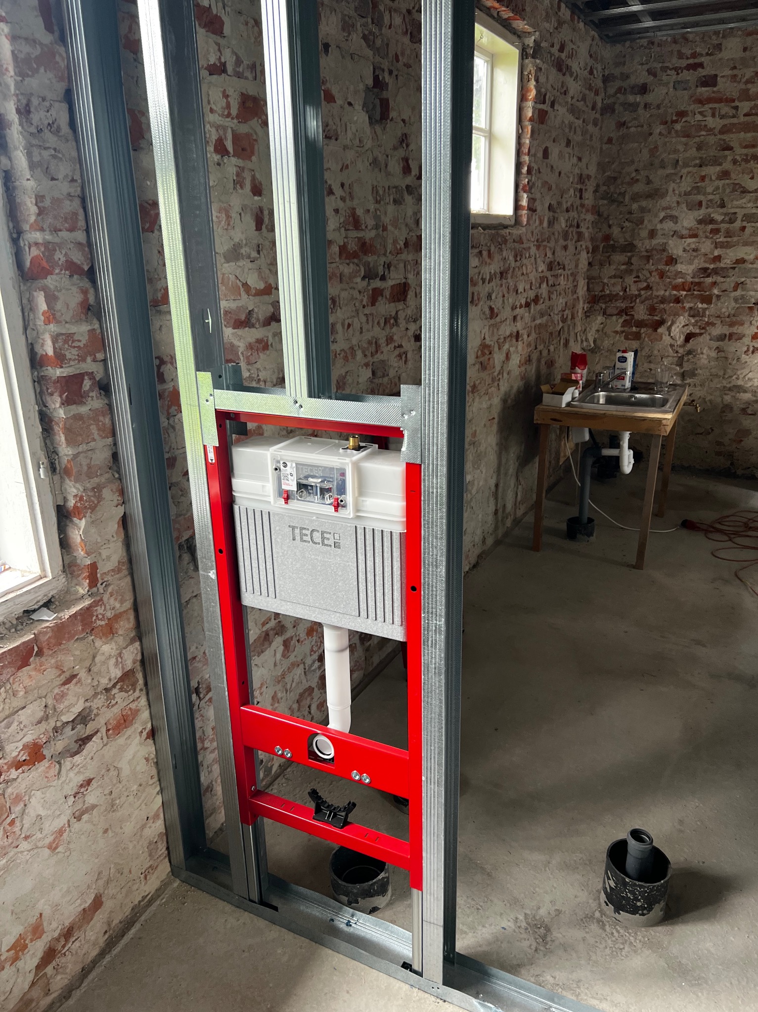 Stelaż podtynkowy TECE zamontowany na metalowej konstrukcji w surowym wnętrzu z ceglaną ścianą. Widoczna instalacja wodno-kanalizacyjna i okno w tle.