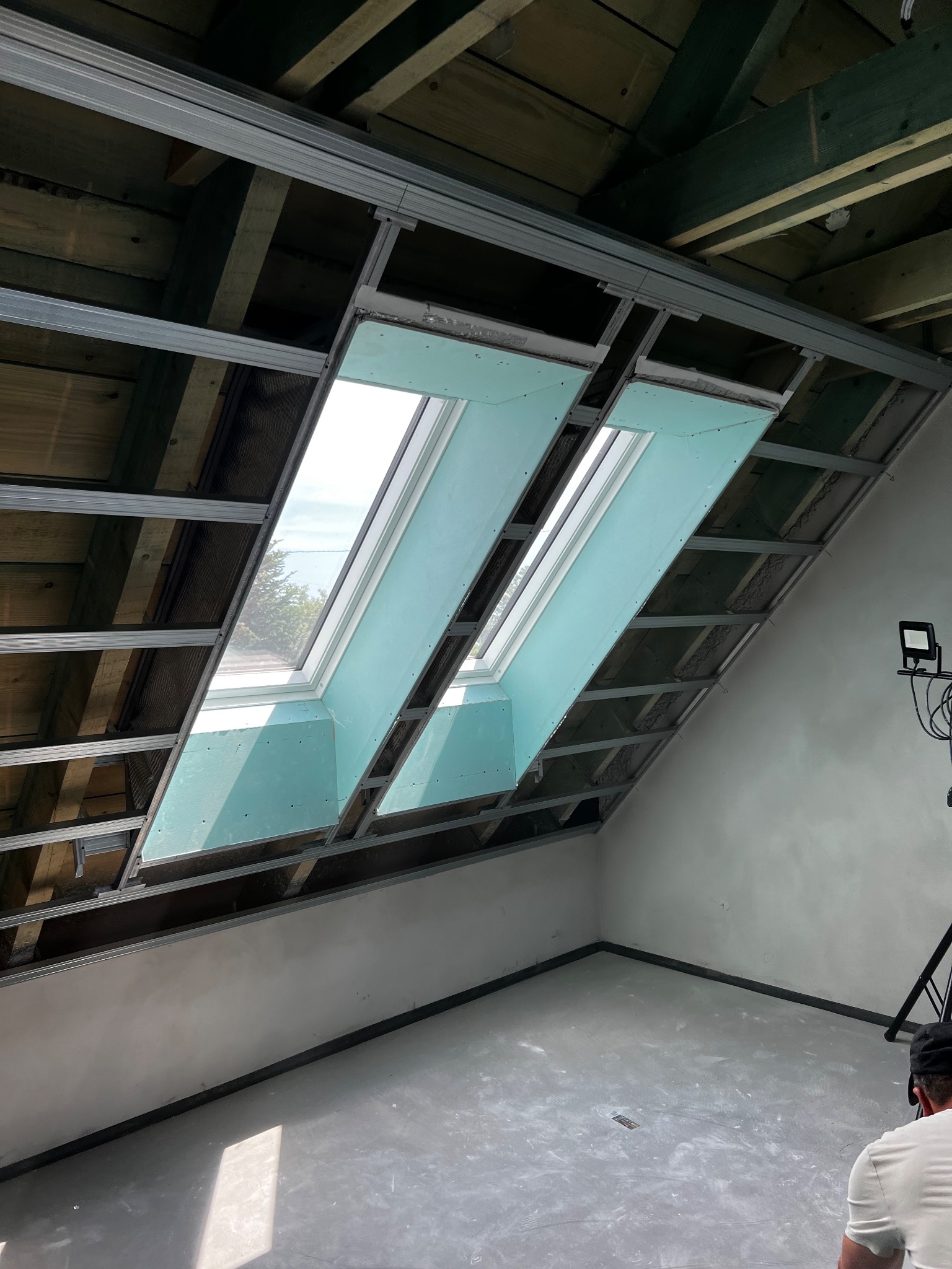 Skośny sufit z dwoma oknami dachowymi w trakcie zabudowy płytami gipsowo-kartonowymi na metalowym stelażu. Widoczne drewniane elementy konstrukcyjne dachu.