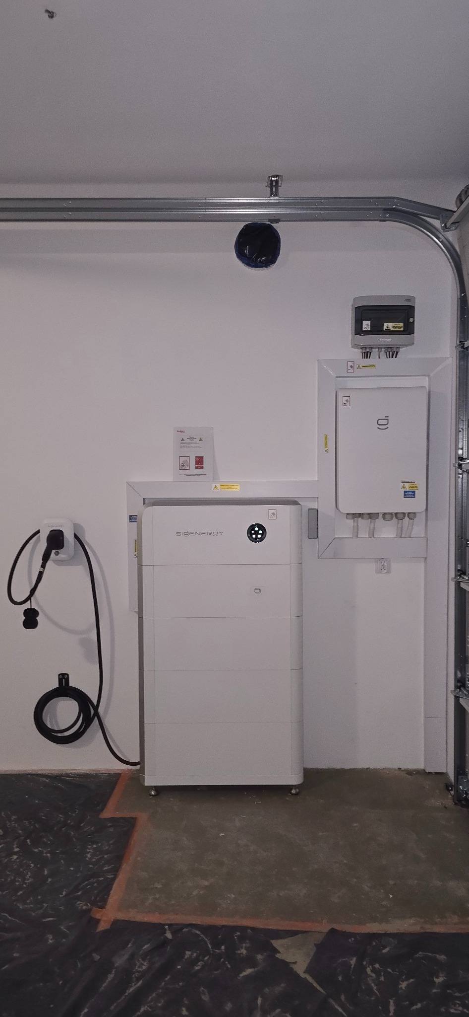 Biały akumulator energii z podłączonym kablem i gniazdkiem na ścianie, obok skrzynka elektryczna. Widoczne elementy instalacji w garażu.