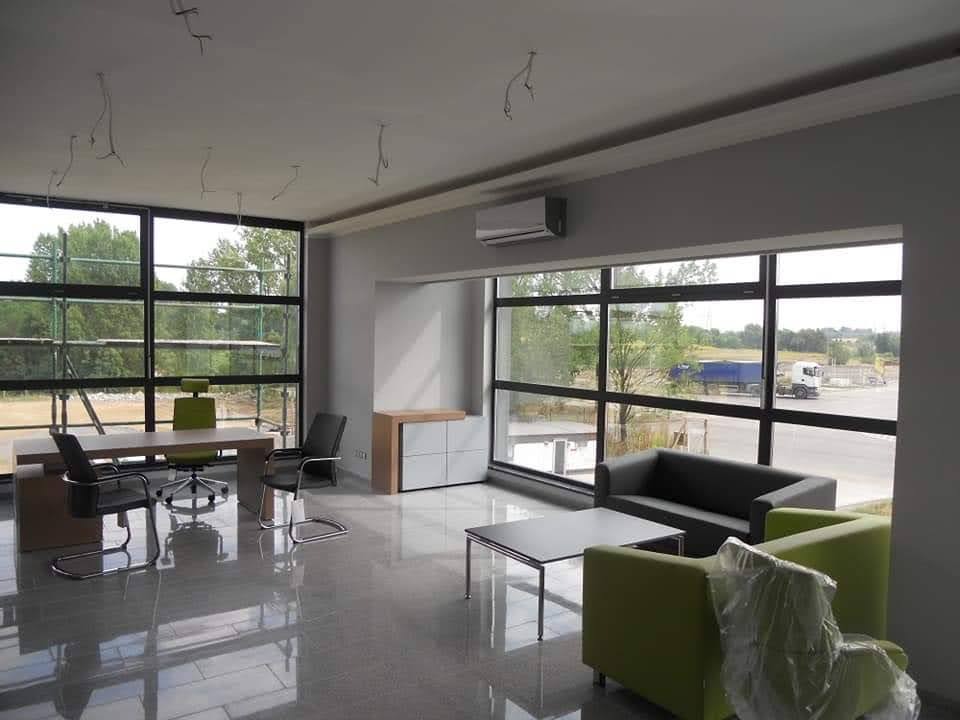 Jasne biuro po remoncie: duże okna z widokiem na zewnątrz, nowoczesne meble biurowe, klimatyzator na ścianie, połyskująca podłoga. Widoczne niedokończone prace na suficie.