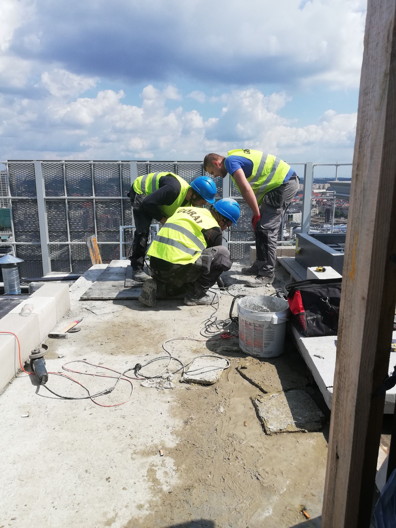 Trzech robotników w kaskach i kamizelkach na dachu budynku podczas prac budowlanych. Widoczne narzędzia, wiadro i elementy konstrukcyjne. W tle panorama miasta i błękitne niebo.