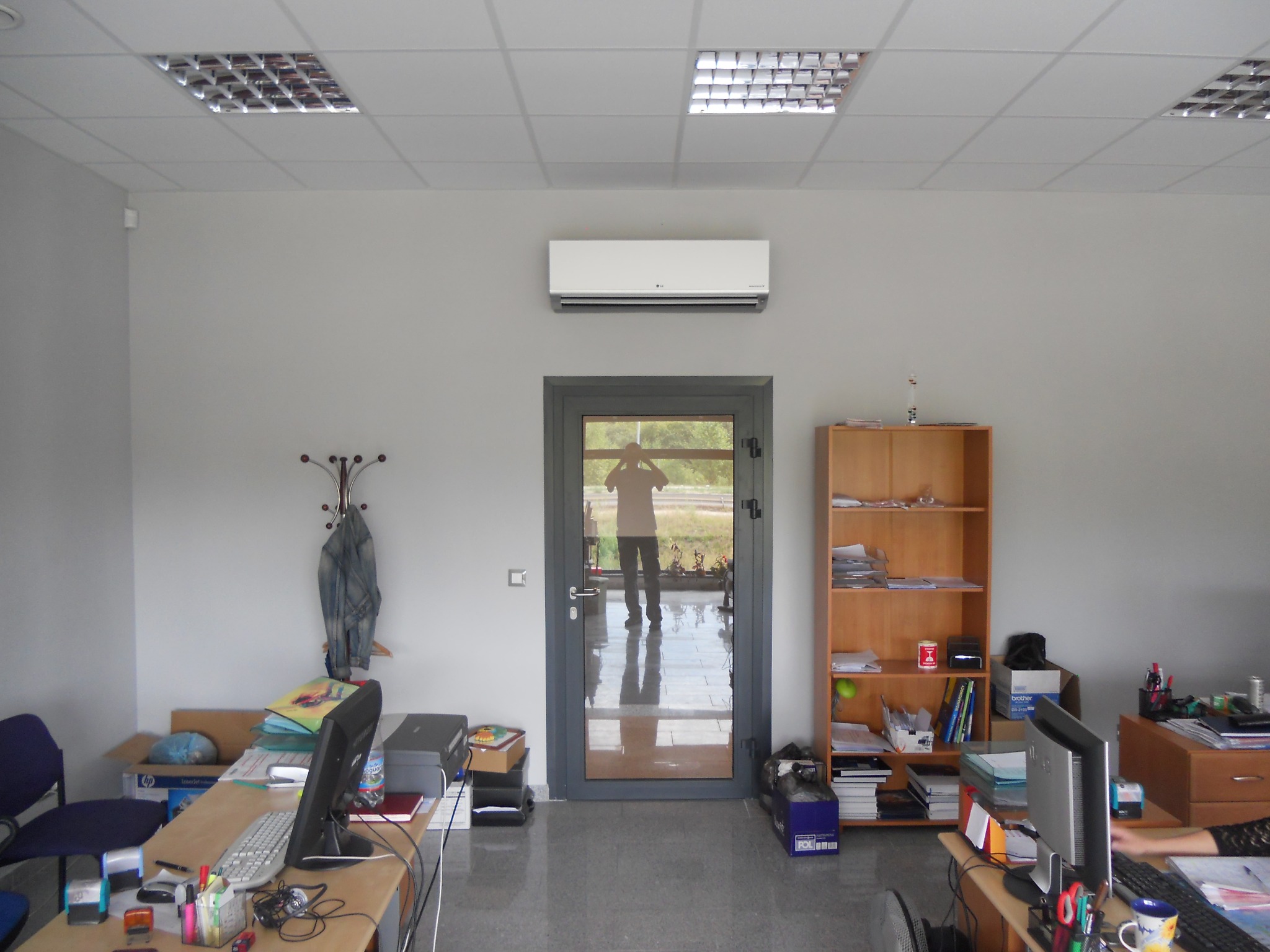 Biały klimatyzator LG zamontowany nad szarymi drzwiami w biurze. Widoczne biurka z komputerami, regał z dokumentami i odblask osoby w drzwiach.