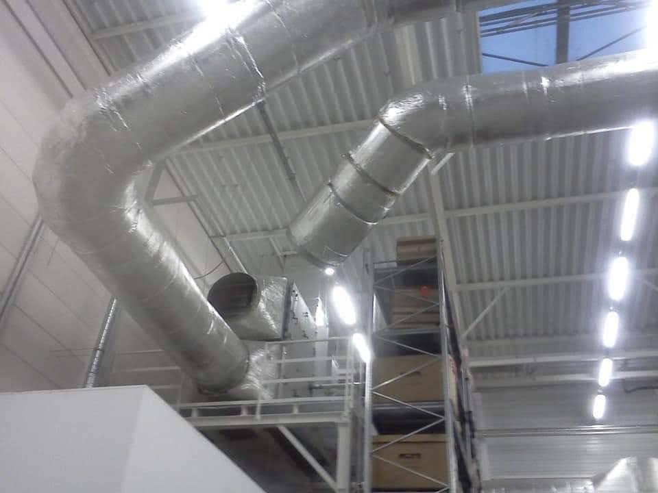 Srebrne, izolowane kanały wentylacyjne zamontowane pod sufitem w hali przemysłowej. Widoczne elementy konstrukcyjne i oświetlenie sufitowe.