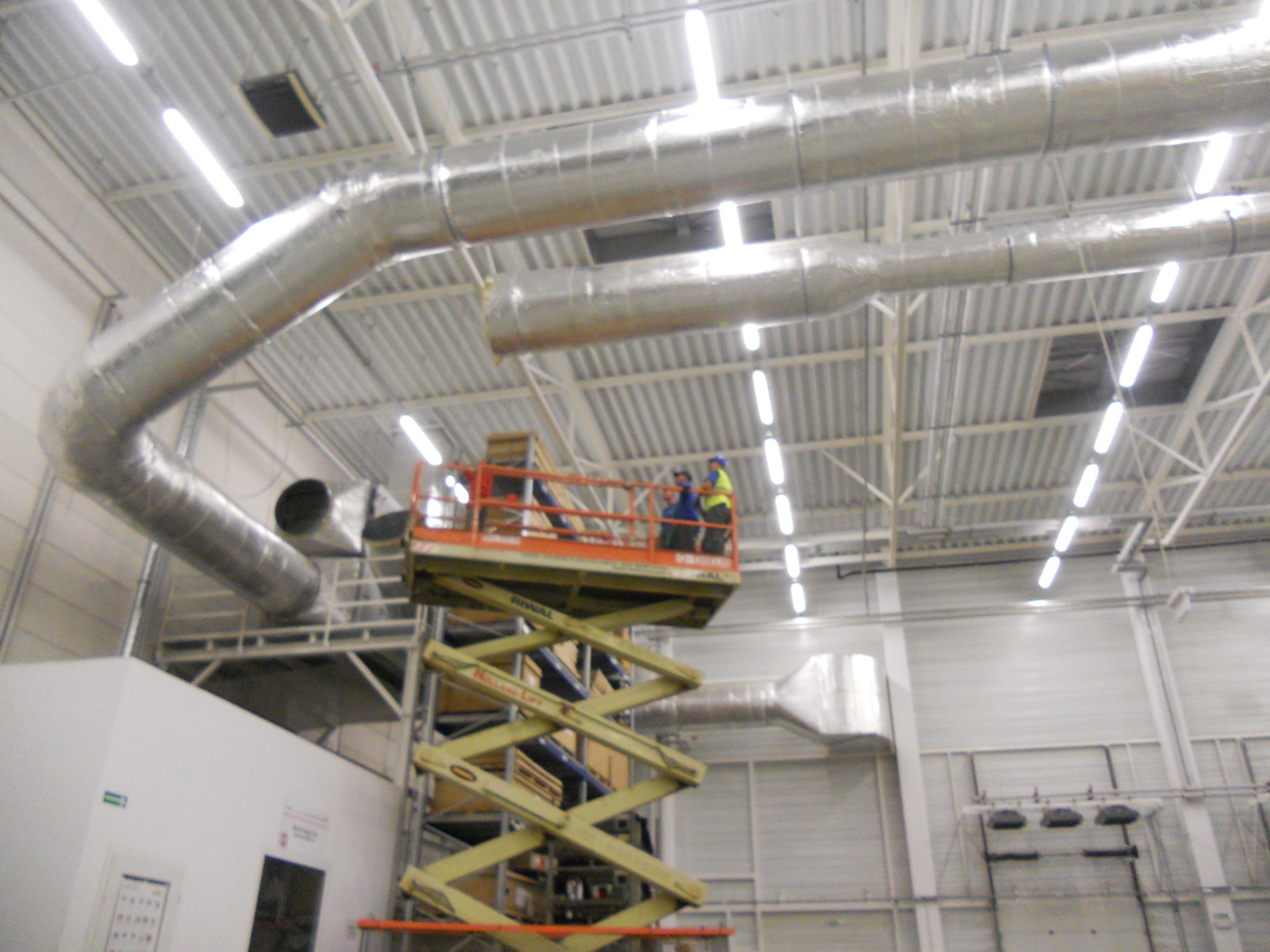Montaż srebrnych rur wentylacyjnych w hali przemysłowej z użyciem podnośnika nożycowego. Widoczni pracownicy w odblaskowych kamizelkach. Oświetlenie jarzeniowe.