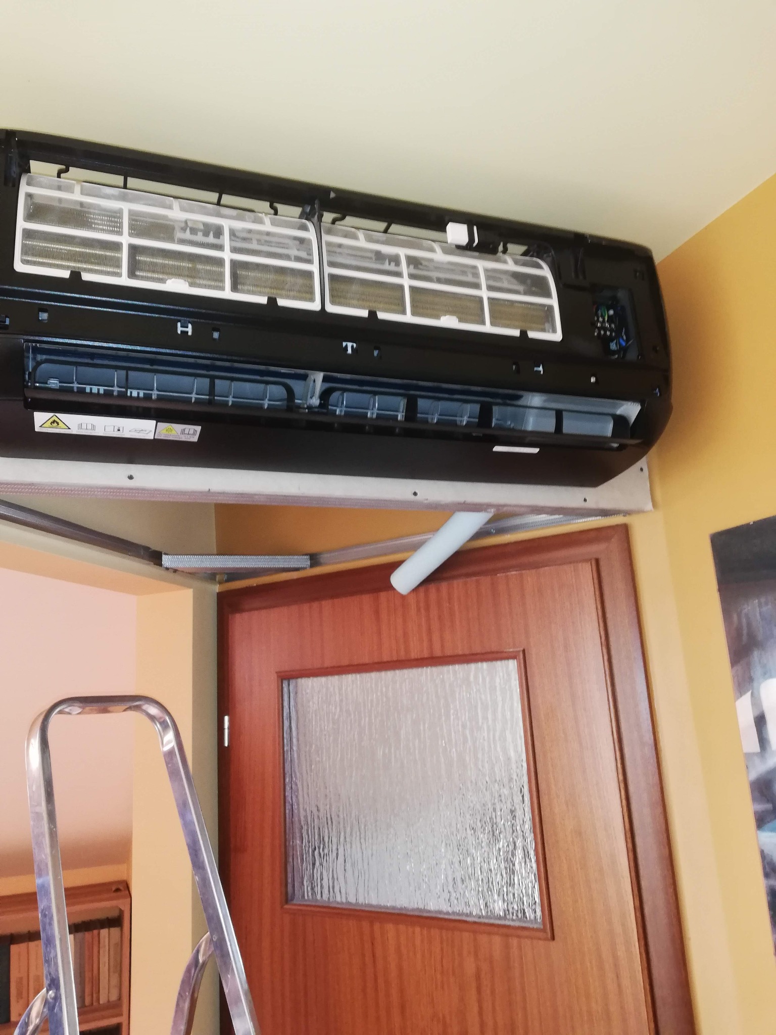 Czarny klimatyzator ścienny z otwartą obudową, widoczne filtry, zamontowany nad drzwiami. W tle drabina i fragment regału z książkami.