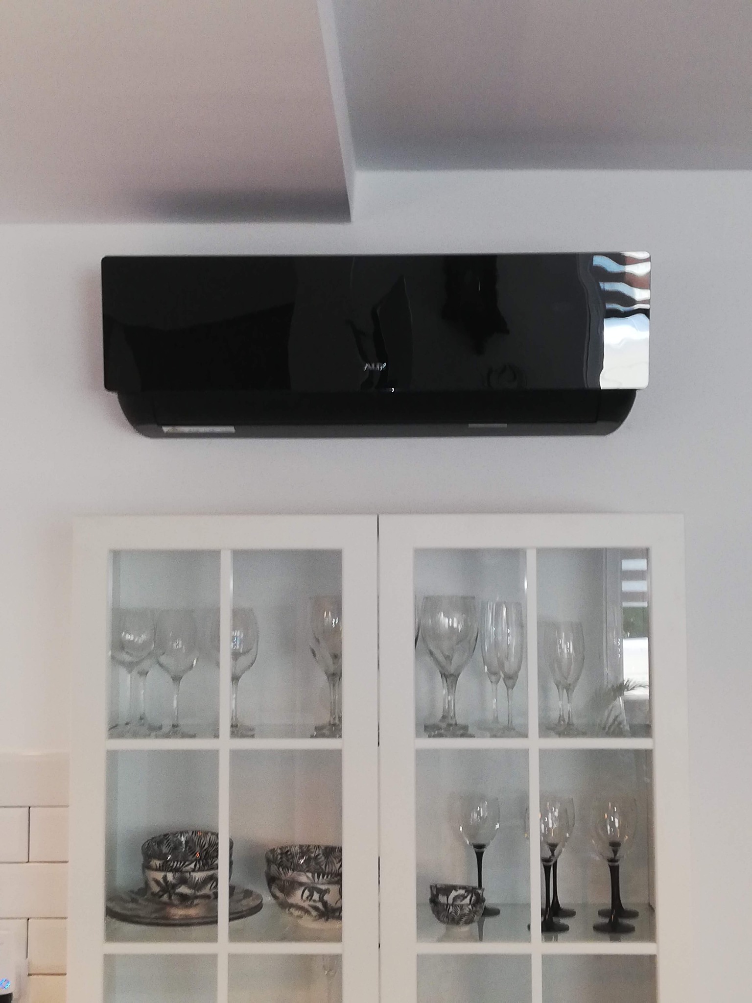 Czarny klimatyzator ścienny nad białą witryną z kieliszkami i miseczkami, nowoczesny design, jasne wnętrze.