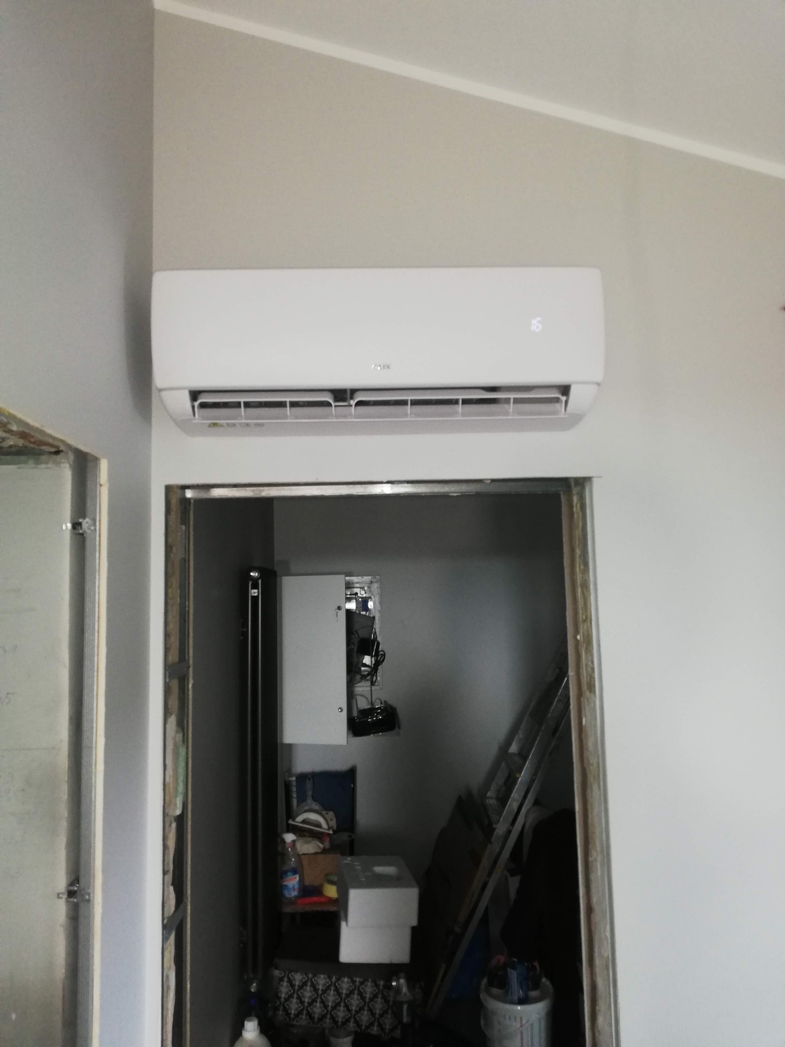 Biały klimatyzator ścienny zamontowany nad futryną drzwiową w szarym pomieszczeniu. Widoczne elementy instalacji w tle.