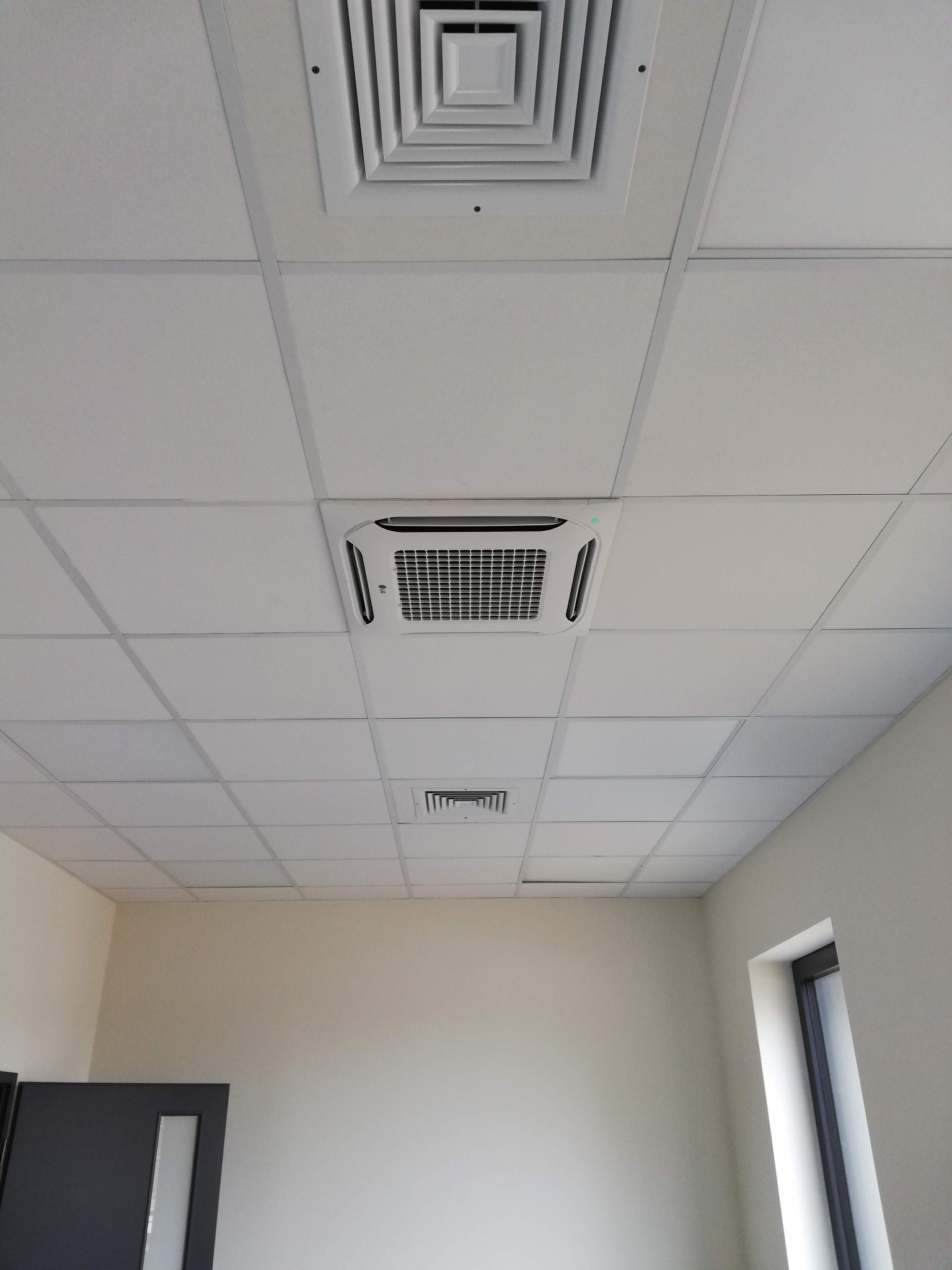 Kasetonowa klimatyzacja sufitowa LG w biurze, widok z dołu. Kratki wentylacyjne w suficie podwieszanym. Minimalistyczny design, neutralne kolory ścian i sufitu.