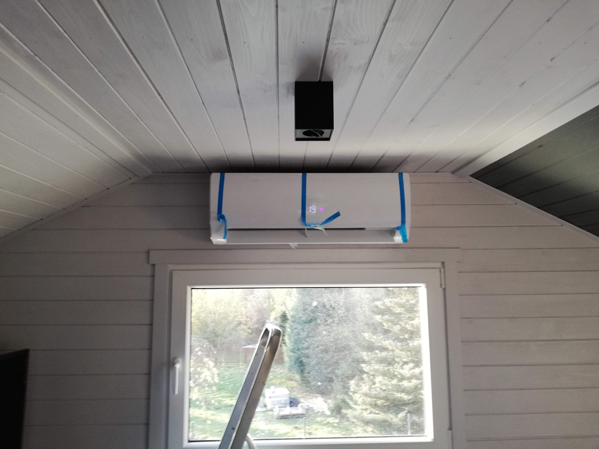 Biały klimatyzator z niebieską folią ochronną zamontowany nad oknem w pomieszczeniu wykończonym boazerią. Widoczna drabina i czarna lampa sufitowa.