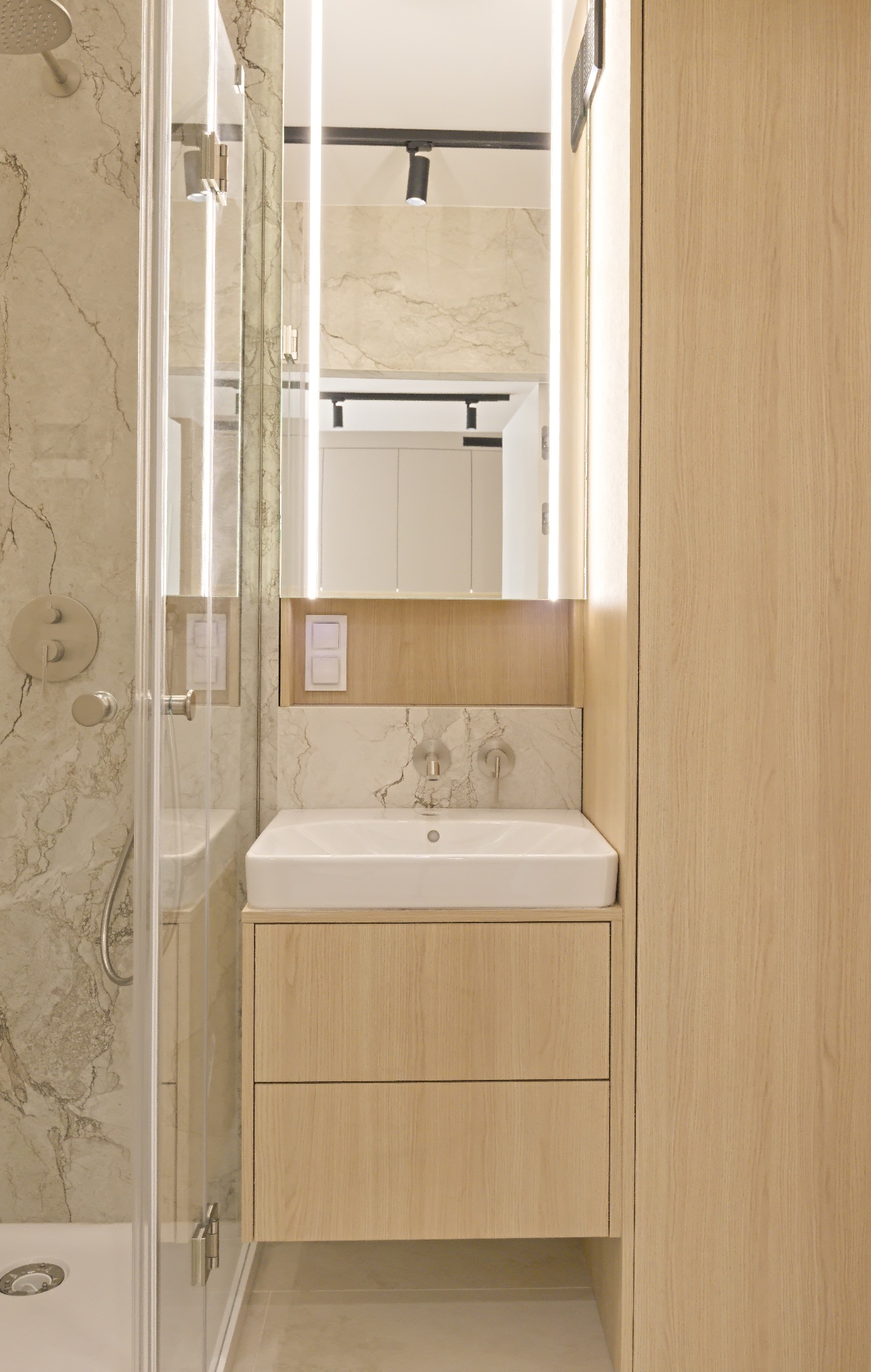 Jasna łazienka z kabiną prysznicową, umywalką nablatową na szafce z dwoma szufladami i podświetlanym lustrem. Marmurowe płytki na ścianach.